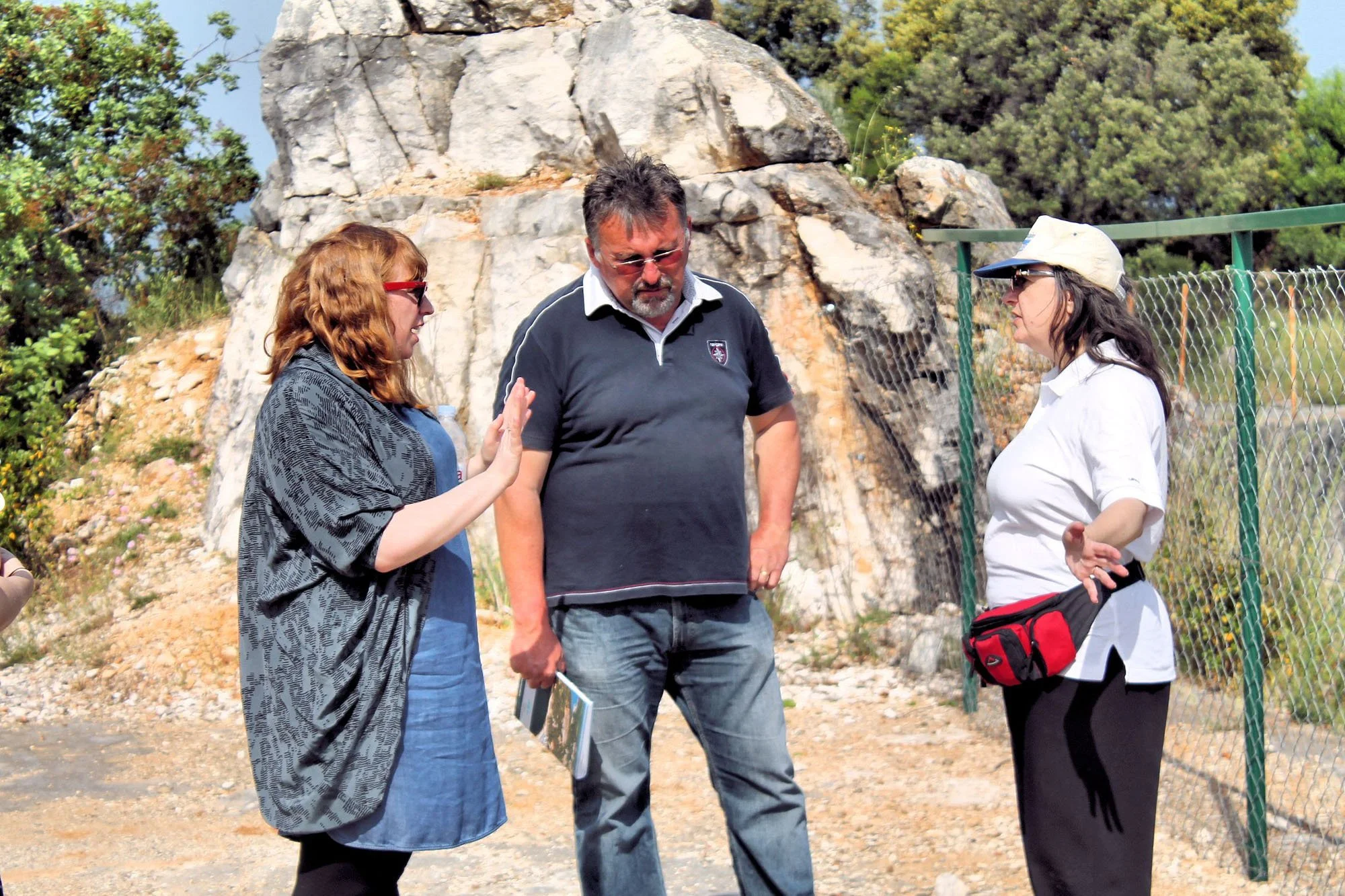 Three people standing outdoors near a rock formation and a chain-link fence, engaged in conversation on a sunny day.