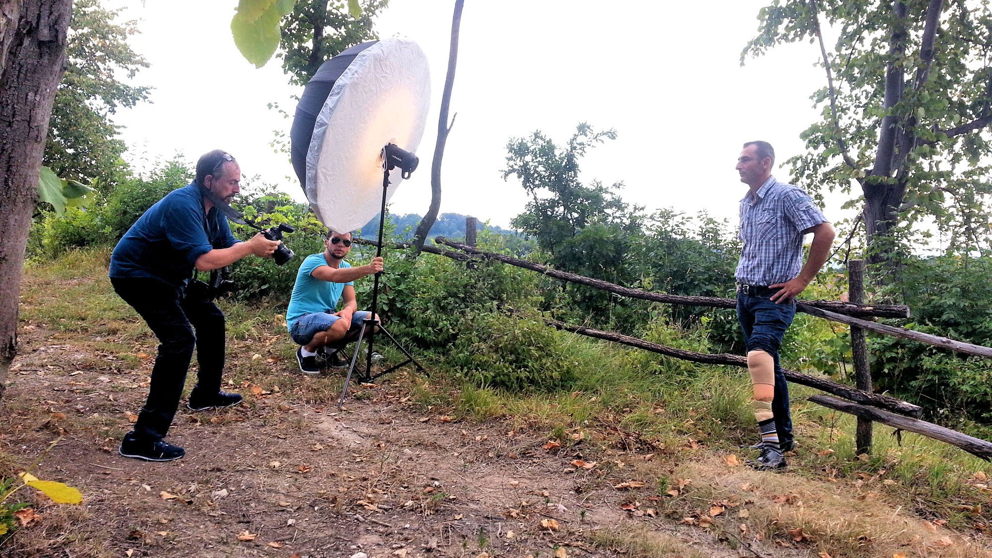 A film crew setting up for a photo shoot outdoors in a wooded area. One person is adjusting a camera, another is holding a large lighting umbrella, and a third person is posing as the subject.