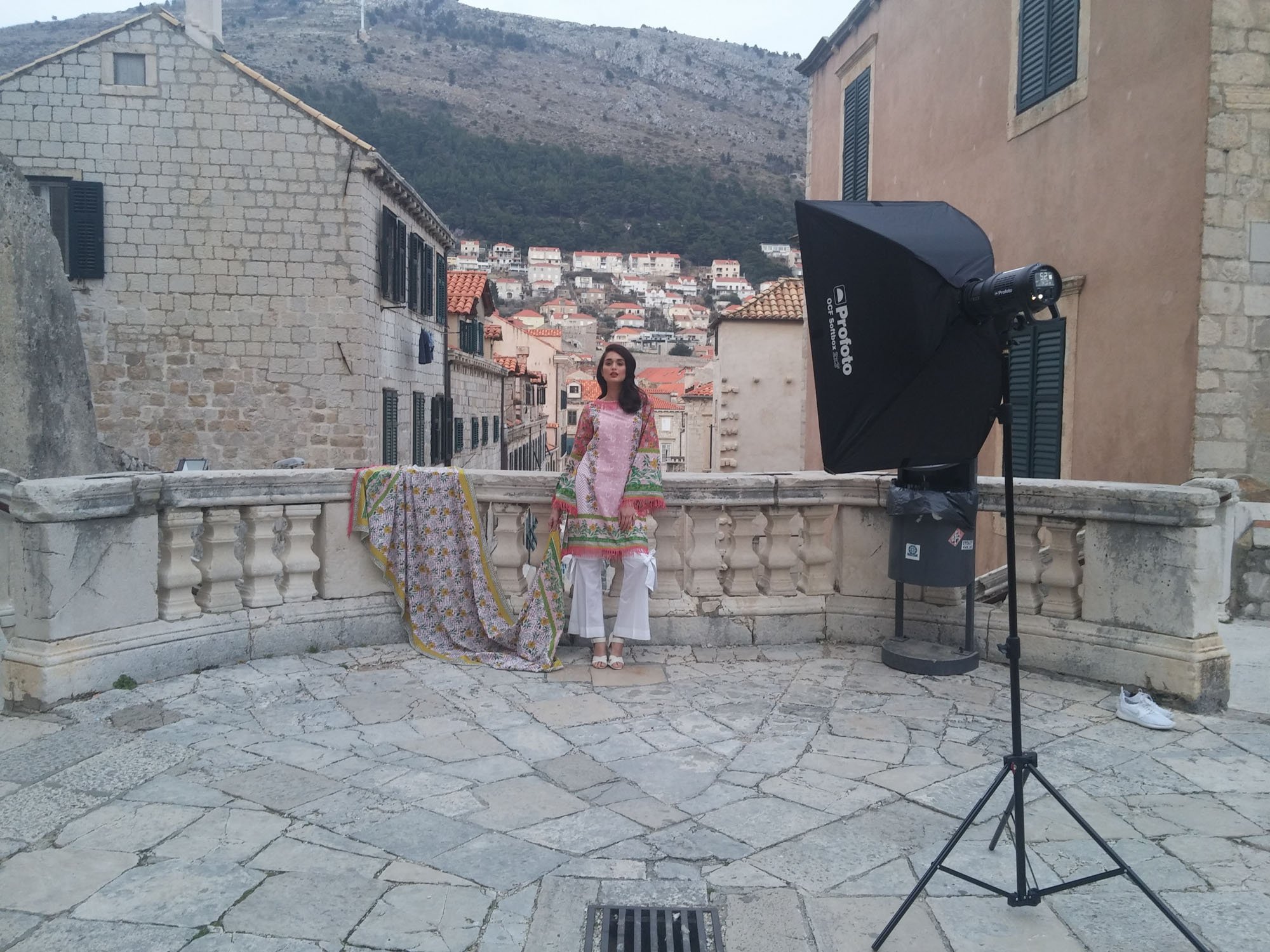 A woman in colorful traditional attire stands on a stone balcony with a mountain and hillside houses in the background, near a photography studio setup including a large softbox and lighting equipment.