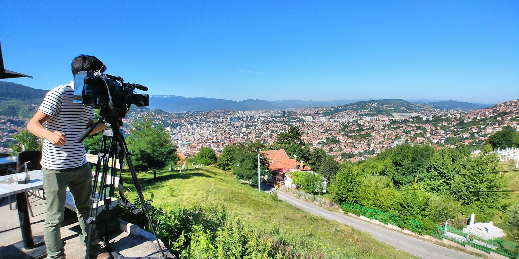 Person filming a cityscape from a hillside, with numerous houses, trees, and mountains in the background under a clear blue sky.