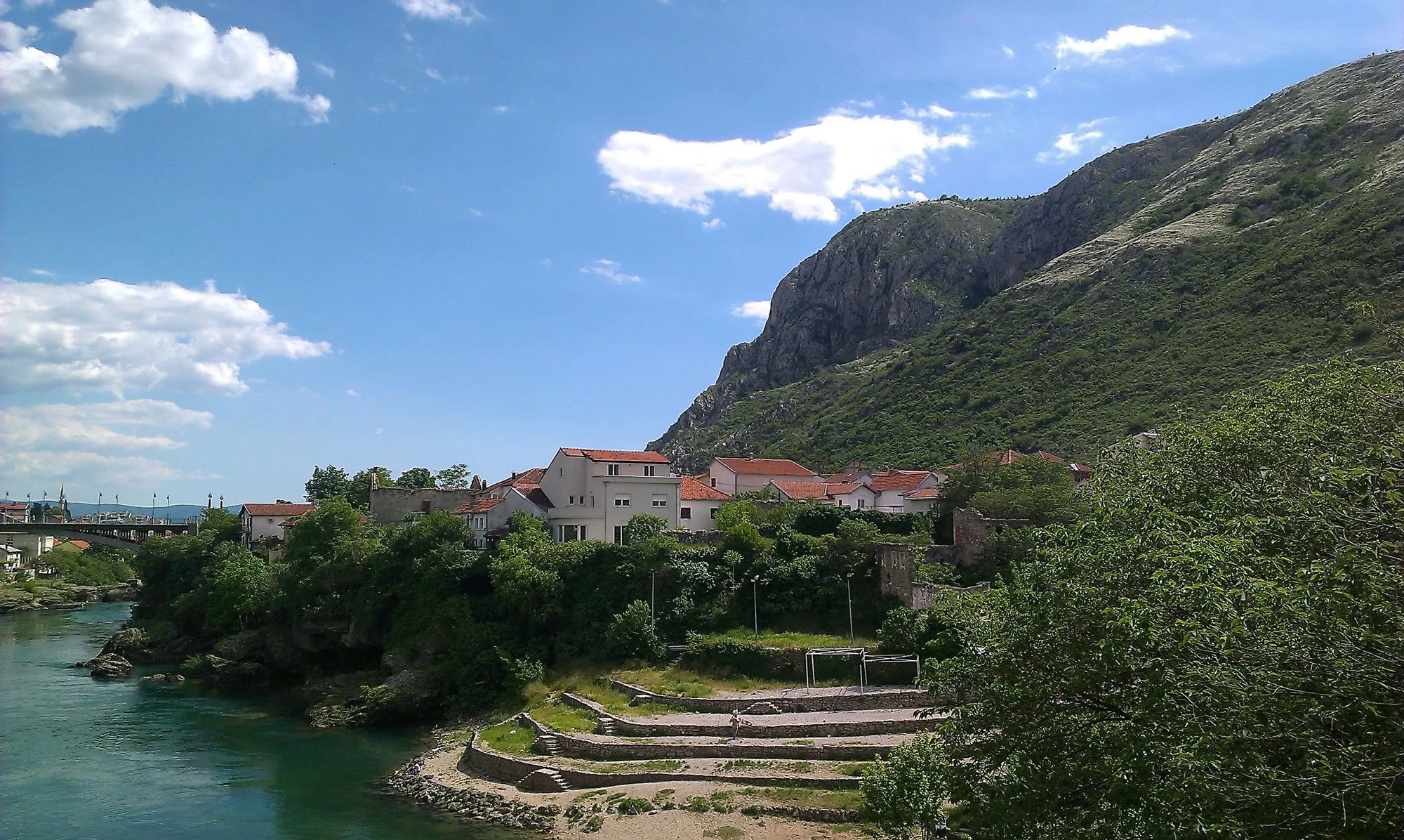 A scenic view of a small town with white buildings and red roofs at the base of a green mountain, next to a river under a partly cloudy blue sky.