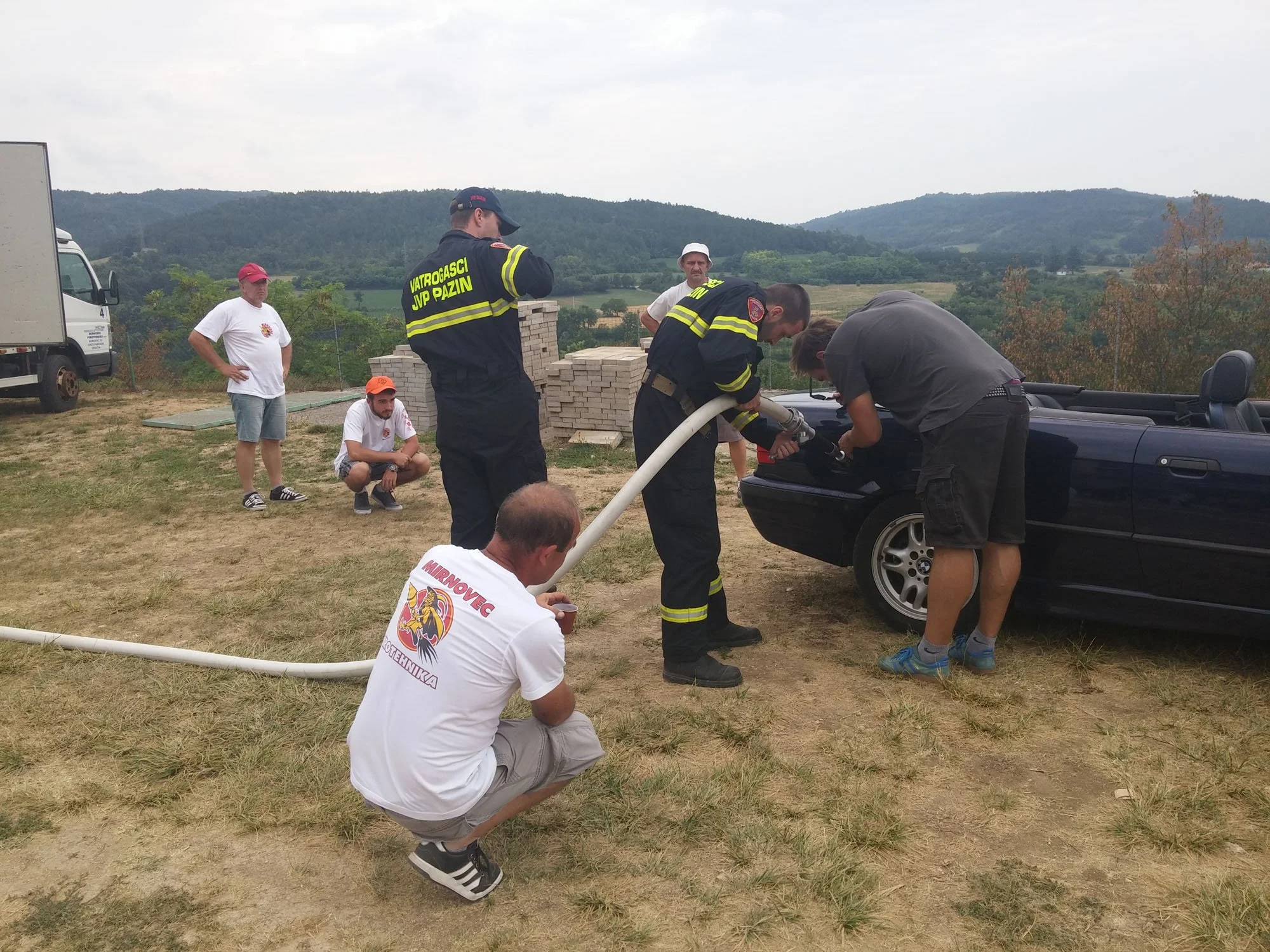 Firefighters and rescue workers at an outdoor scene, attending to an emergency involving a black convertible car and a hose, with mountains and cloudy sky in the background.