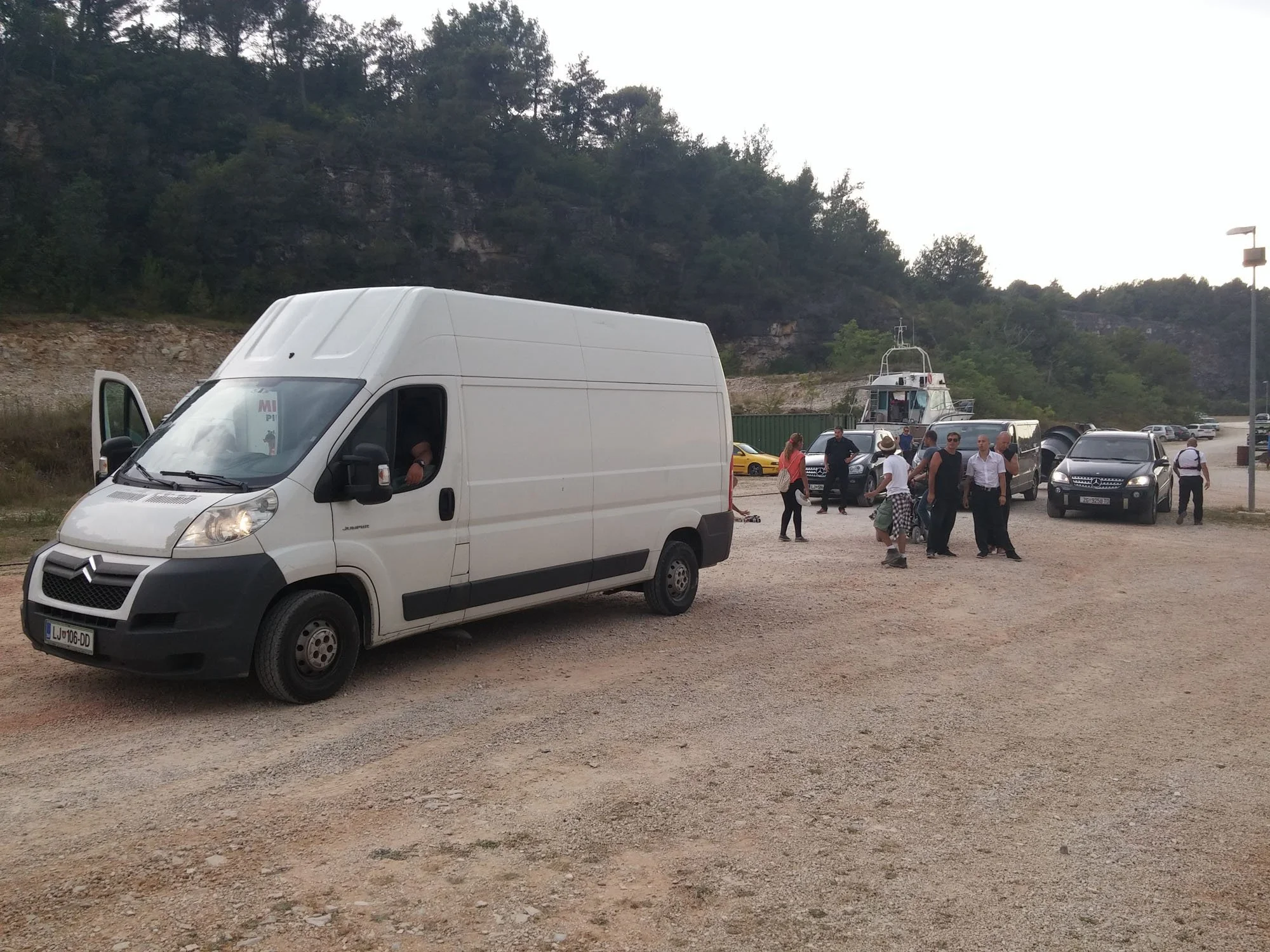A white delivery van parked in a gravel lot with an open door, a group of people walking and standing nearby, a boat in the background, and a hillside with trees