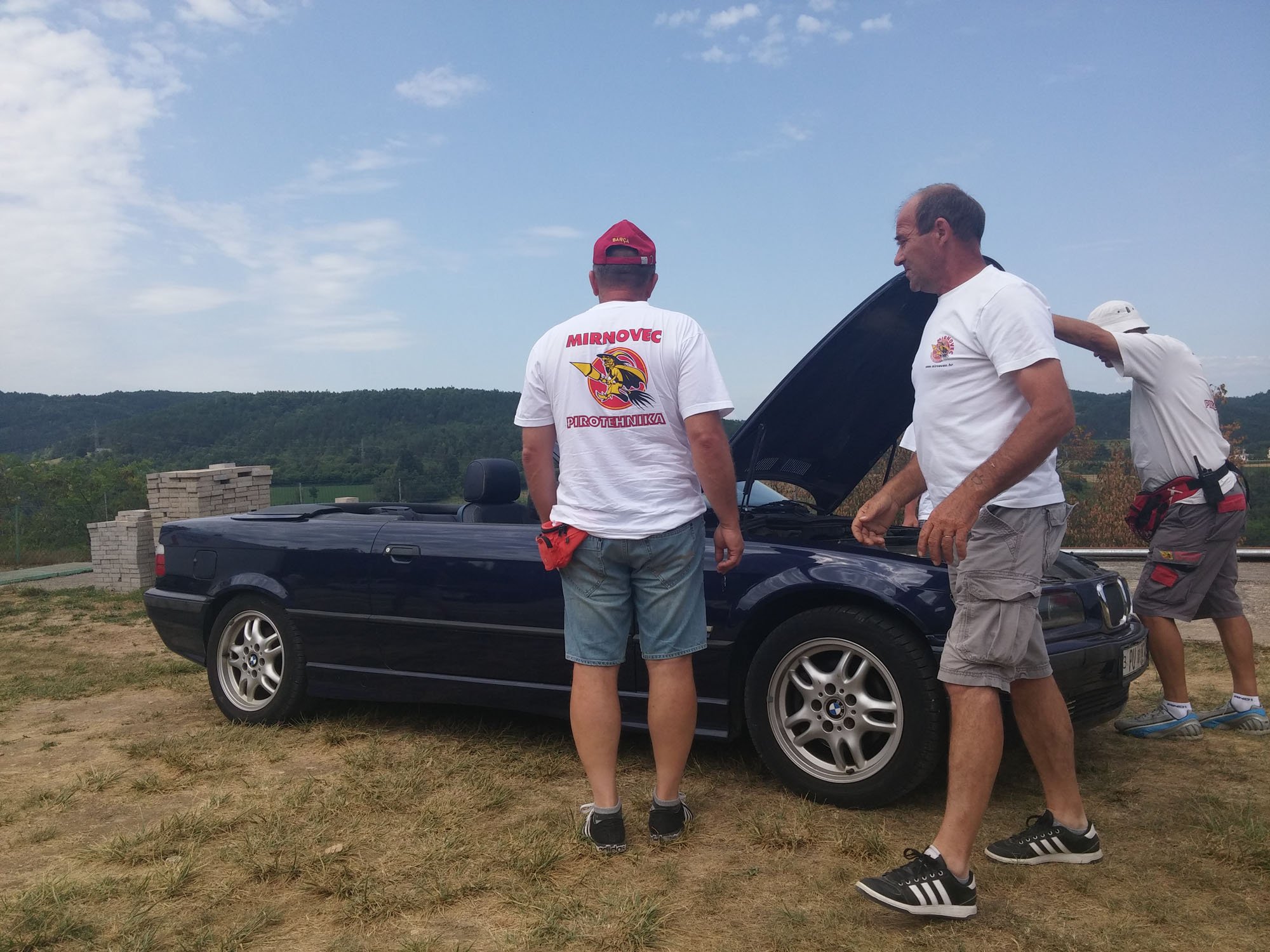 Three men stand beside a black convertible car outdoors, with the hood open, inspecting it. The man in the middle wears a red cap and a white T-shirt with a colorful lion and text. The man on the right wears a white T-shirt, and the person on the far