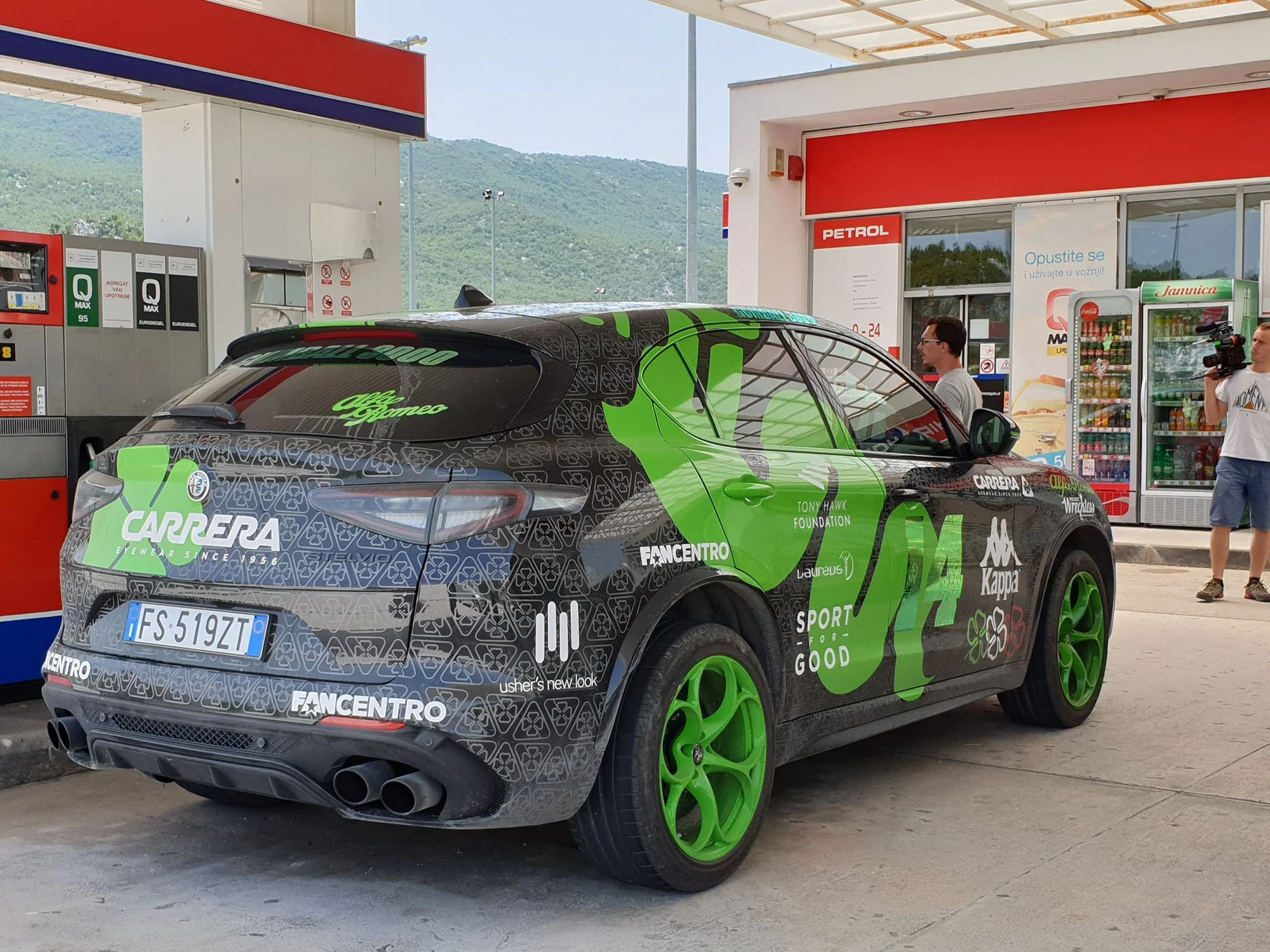 A black and green sports car parked at a gas station with mountains in the background. The car has sponsors' logos and the words 'Carrera' and 'Fancentro' on it, and there is a man with glasses and a camera standing near the car.