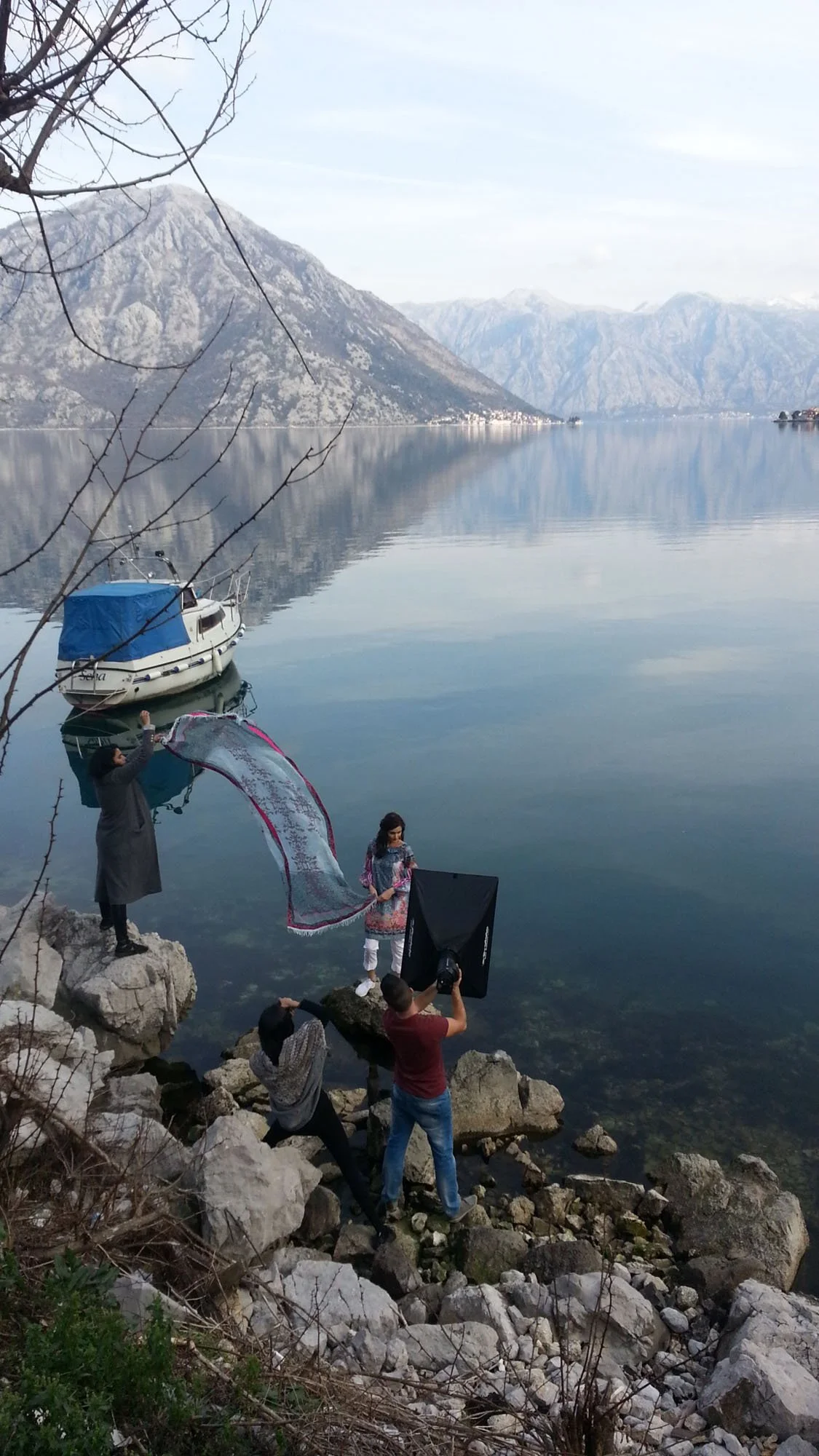 A photoshoot setup by a lake with mountains in the background. A woman in traditional dress is posing on rocks, while photographers and assistants are adjusting lighting and equipment.