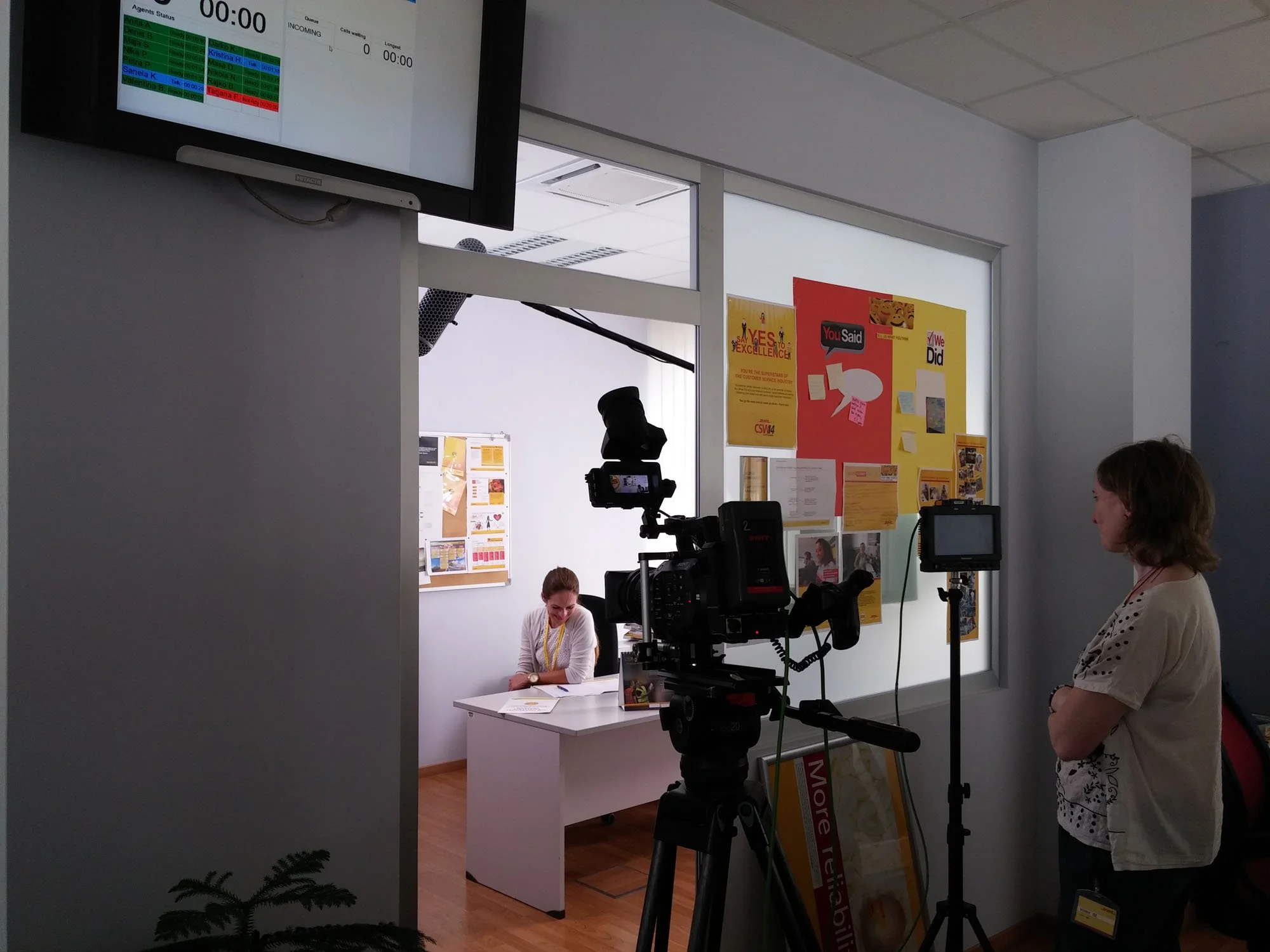 A newsroom or interview setting with a person sitting at a white desk, smiling, and wearing a necklace, with a woman standing nearby. There are cameras and filming equipment directed at the woman. The background features a wall with posters and infor