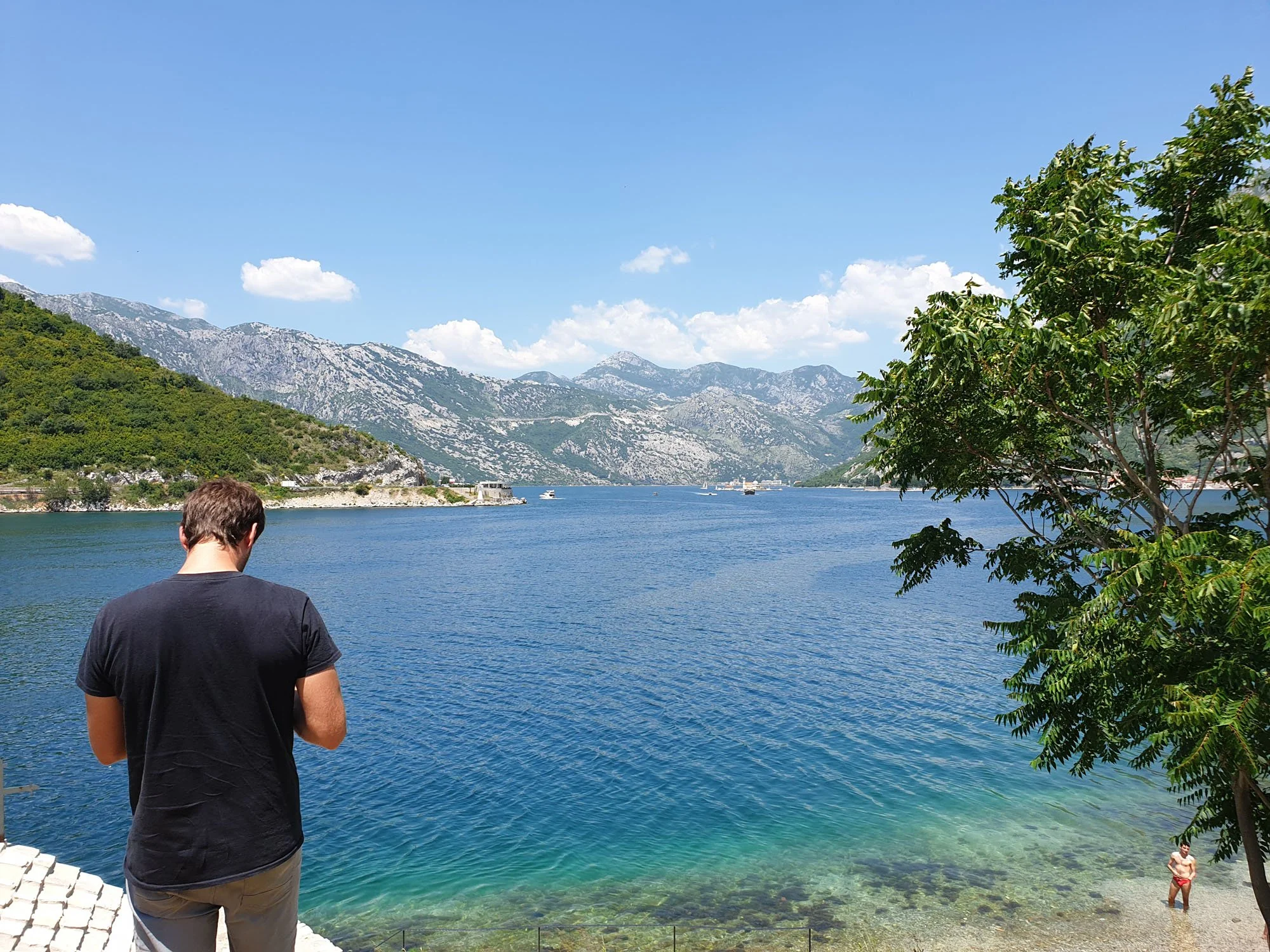 A man in a black shirt is standing near a body of water, with a person in red swimming trunks in the water. In the background, there are mountains and a partly cloudy blue sky, with trees on the right side of the image.