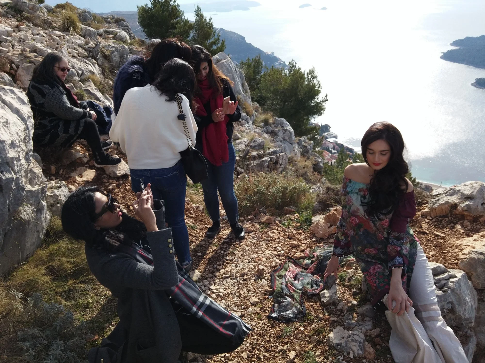 A group of women are gathered outdoors on rocky terrain overlooking a large body of water with hills or islands. Some women are sitting on rocks, and one woman appears to be in the process of adjusting her clothing or preparing for a photo. They are 