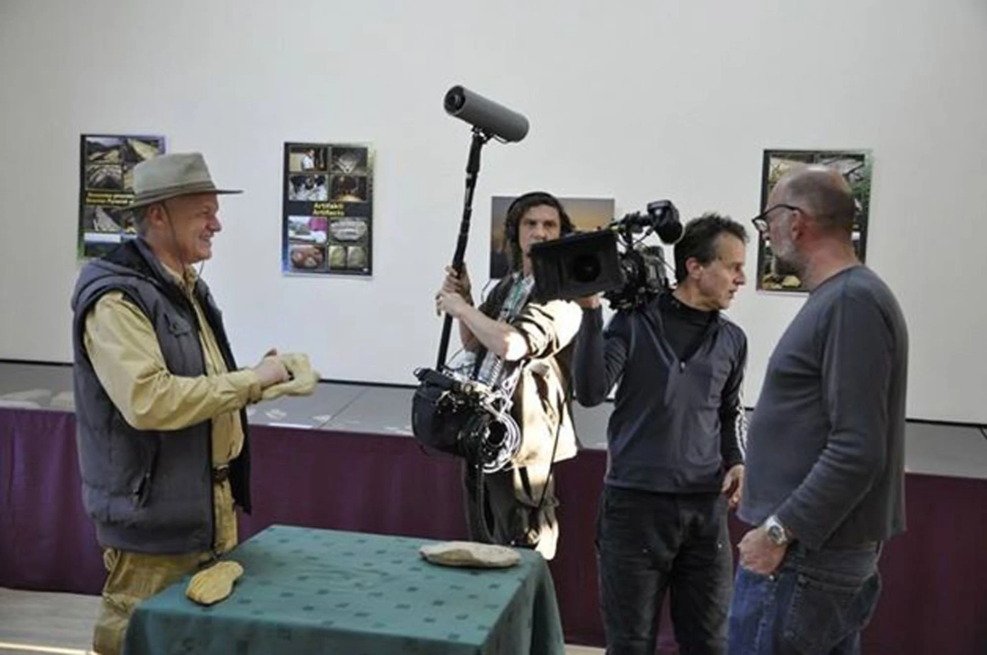 A group of four men engaged in conversation at an indoor event. One man on the left is smiling and holding a sandwich, wearing a beige hat and a vest. Another man in the middle is operating a professional video camera with a boom microphone, while th