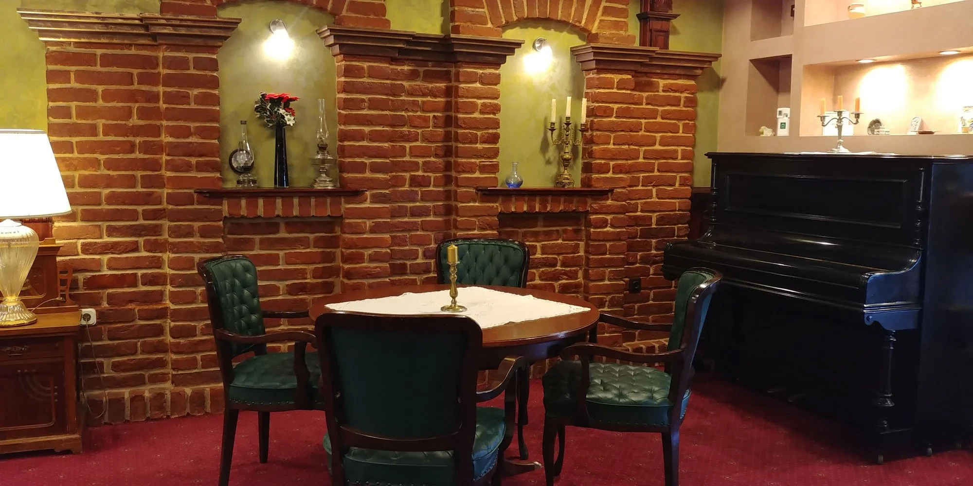 A cozy room with a brick wall, a black upright piano, a round wooden table with a white lace doily and a gold candle holder, green upholstered chairs, a lamp on the side table, and decorative items such as candlesticks, vases, and candles on shelves 