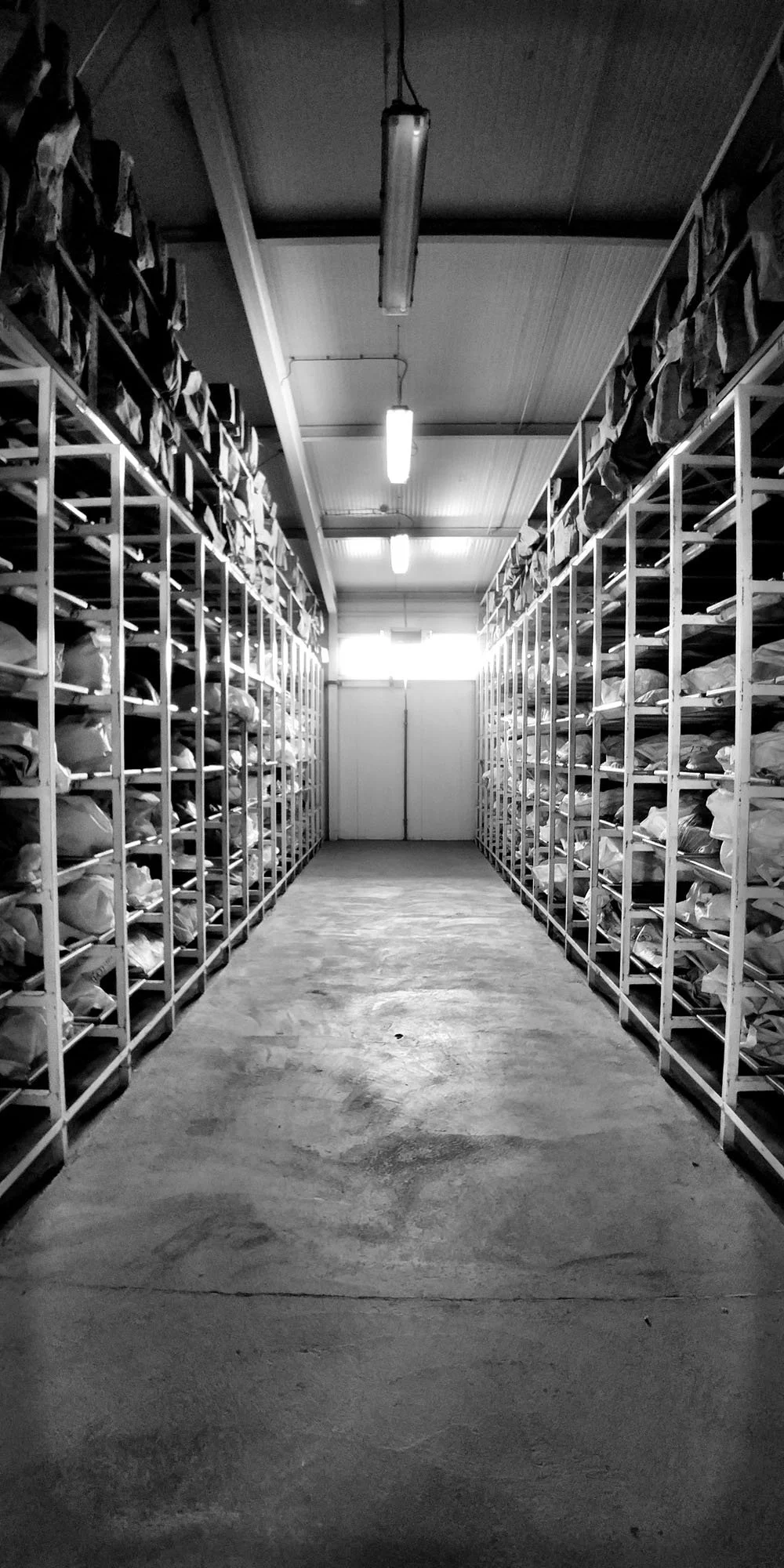 A black and white photo of a storage room with metal shelves on both sides holding numerous bags or packages, and a concrete floor leading to a closed door at the far end.