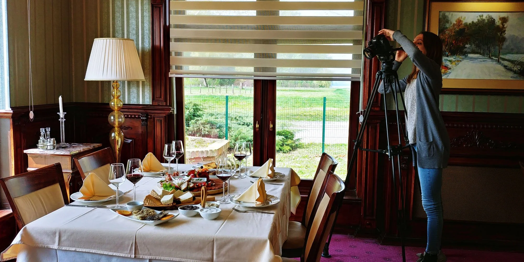 A woman is setting up or adjusting a camera on a tripod in a dining room with a large window overlooking a garden. The round table is set for a meal with wine glasses, plates of food, cheese, and appetizers. The room has wooden paneling and artwork o