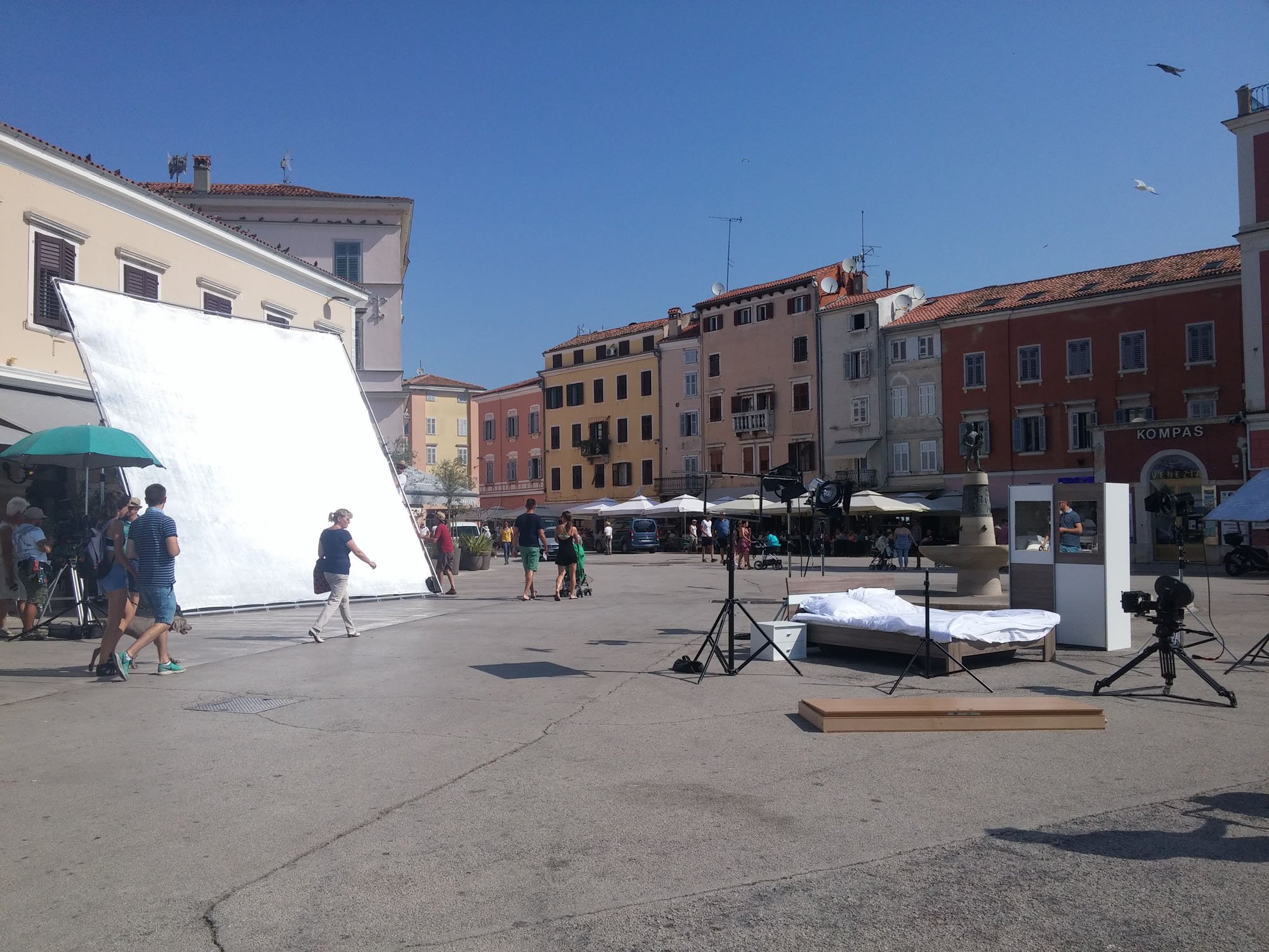 A square with people walking and seated at outdoor cafes, surrounded by colorful buildings, with studio equipment and a backdrop, under a clear blue sky.