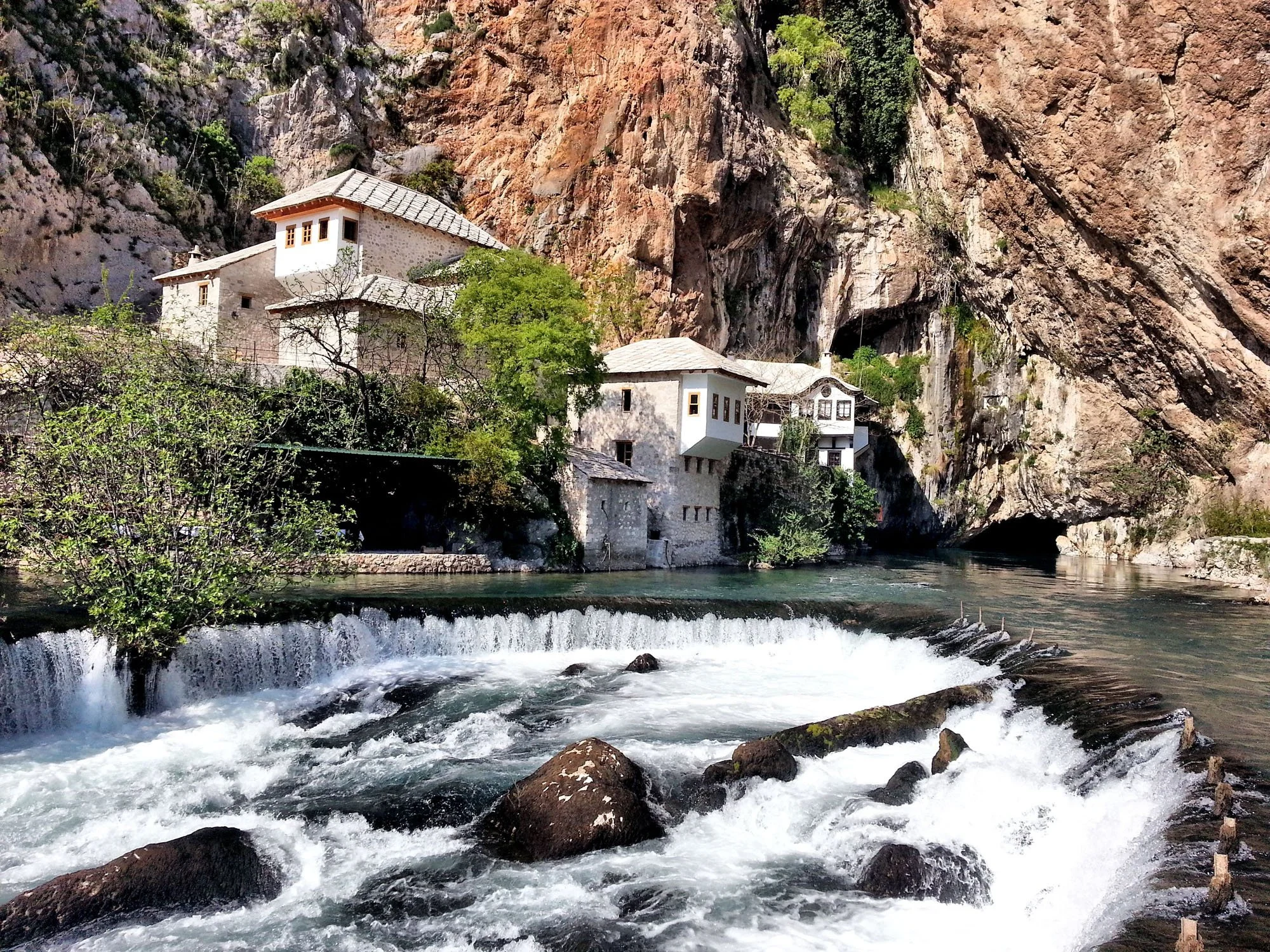 White houses with brown roofs situated on rocky cliffs beside a flowing river, with a large cave entrance visible in the rocky hillside.