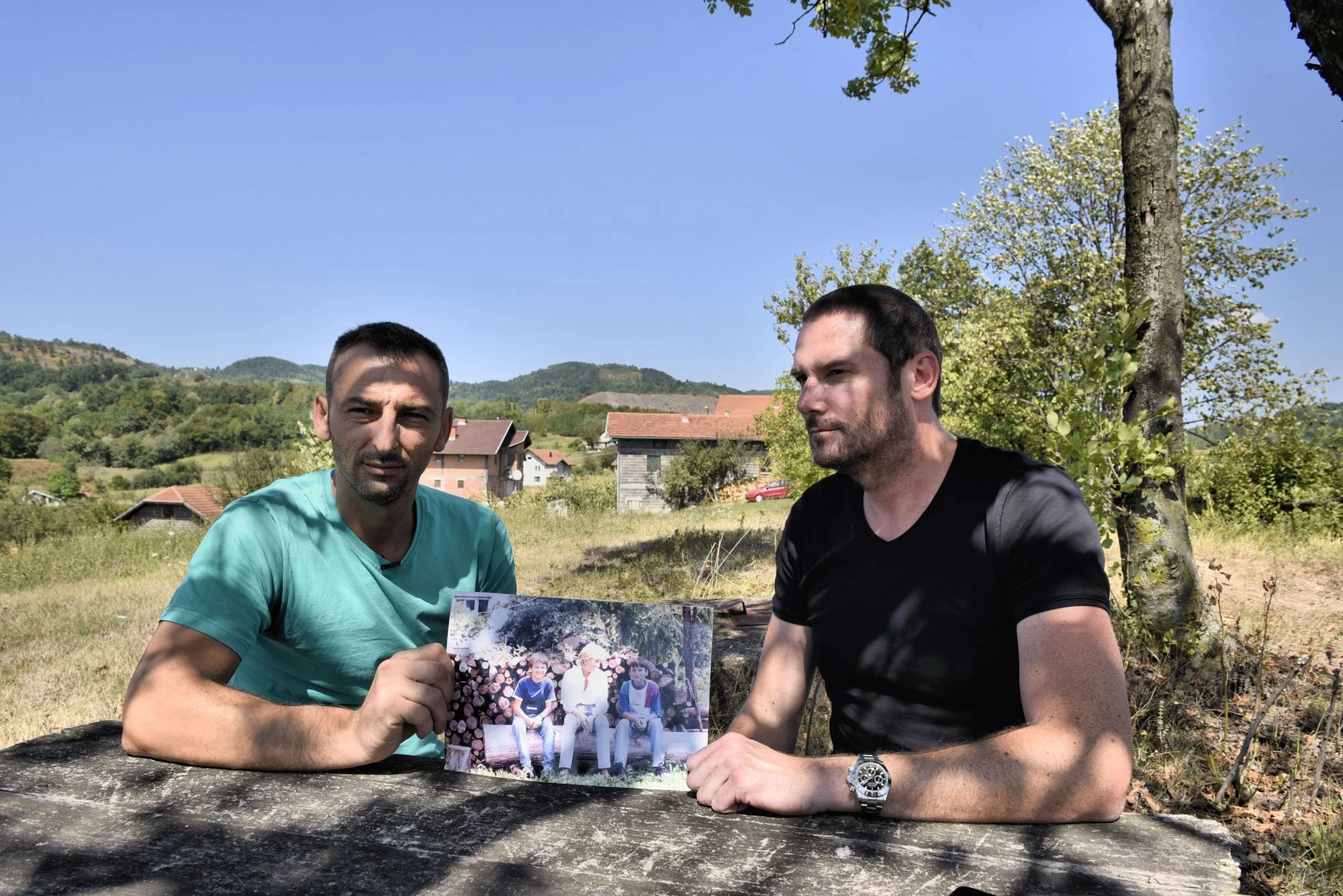 Two men sitting outdoors under a tree, one holding a photo of three people sitting together, with a rural landscape and house roofs in the background on a sunny day.