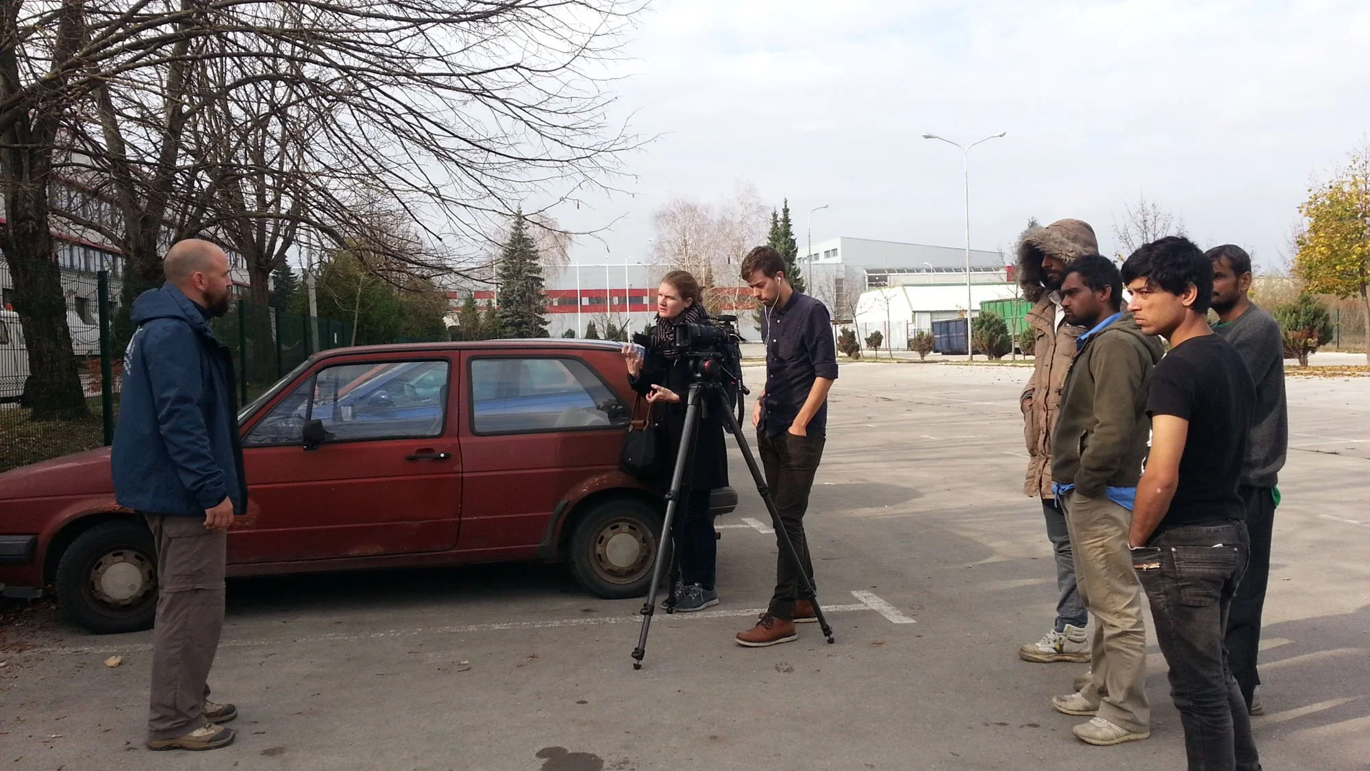 Group of people standing outdoors near a red car in a parking lot, with a person filming them and another person speaking, among winter trees and industrial buildings in the background.