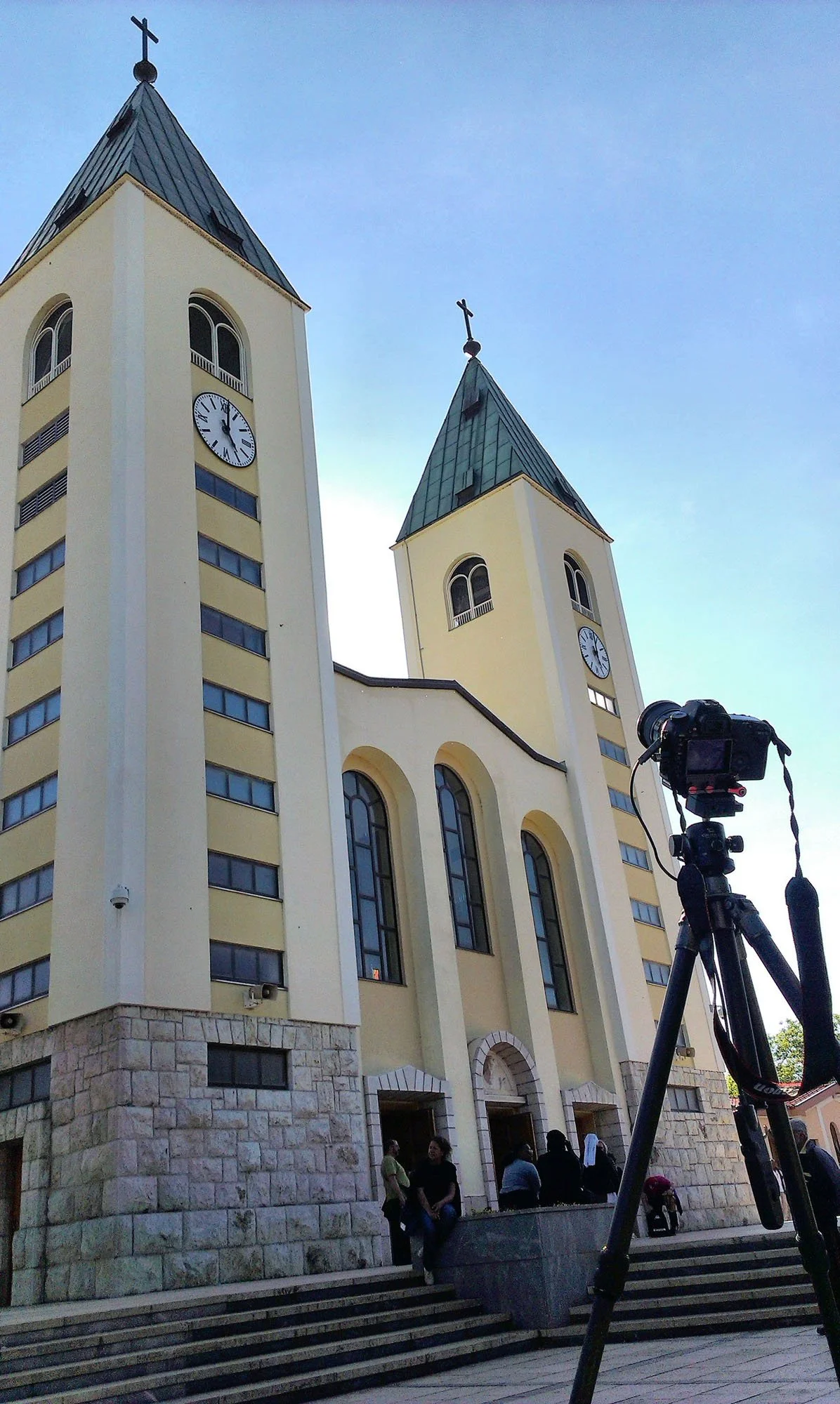 A church with two steeples, clocks on each tower, and people standing and sitting near the entrance. A professional camera on a tripod is in the foreground.
