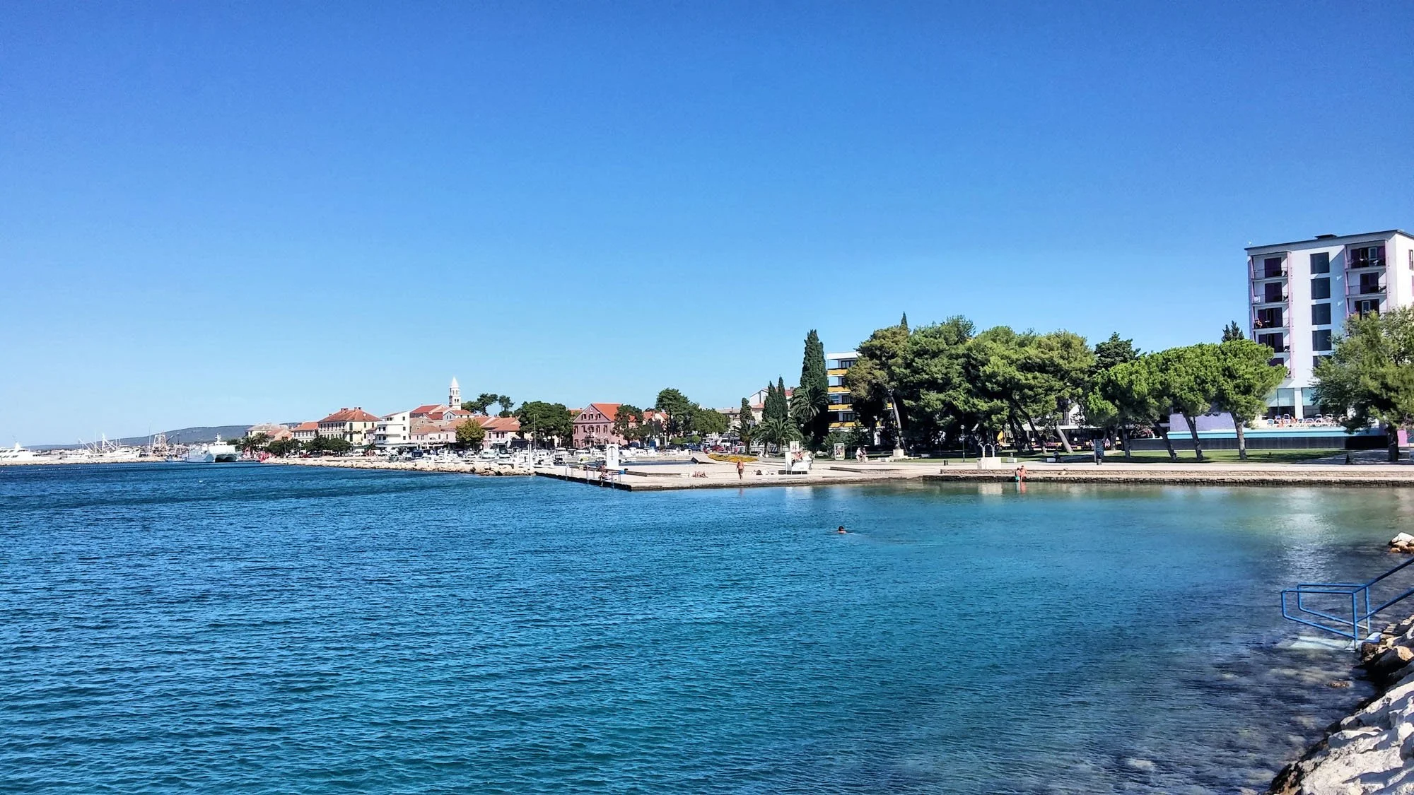 View of a marina with calm blue water, a tree-lined waterfront, and a village with buildings, including a church steeple, under a clear blue sky.