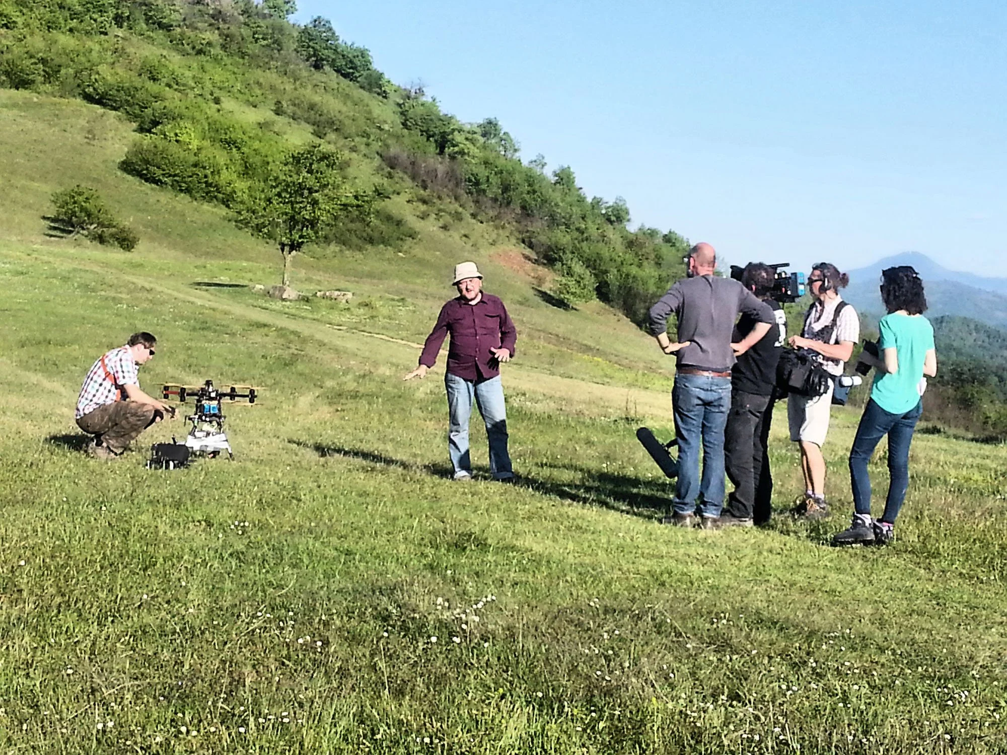 A group of people gathered outdoors on a grassy hillside with mountains in the background. Some are operating cameras, while others are standing or crouching near a drone, with a man in a purple shirt and beige hat talking.