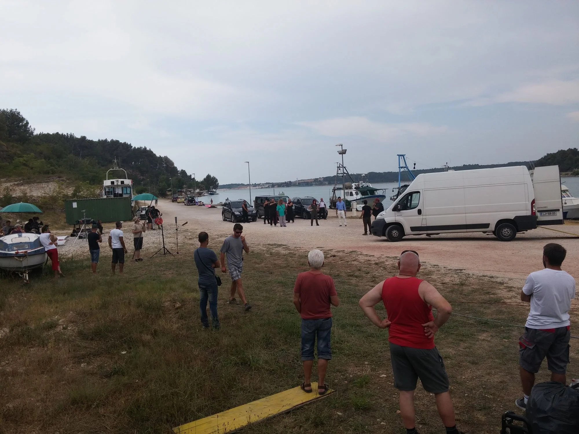 People gathered near boats and cars on a coastline, possibly preparing for or returning from a boat trip, with cloudy skies overhead.