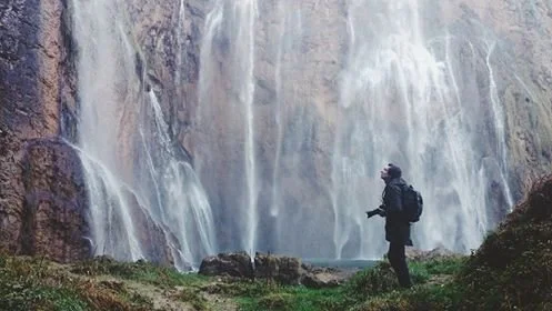 Hiker with backpack standing near a waterfall in a forest.