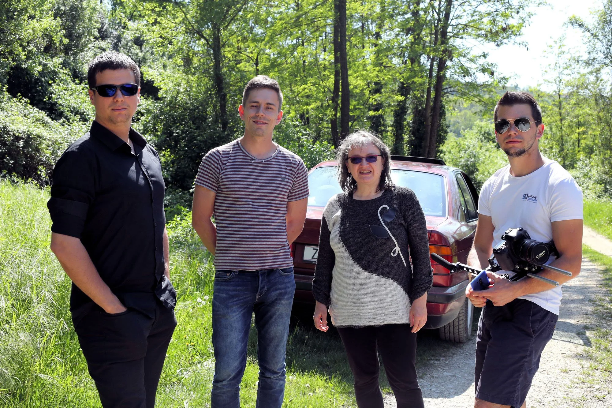 Four people standing outdoors in a wooded area with green foliage and sunlight, two men wearing sunglasses, one woman, and one man holding a professional camera and tripod.