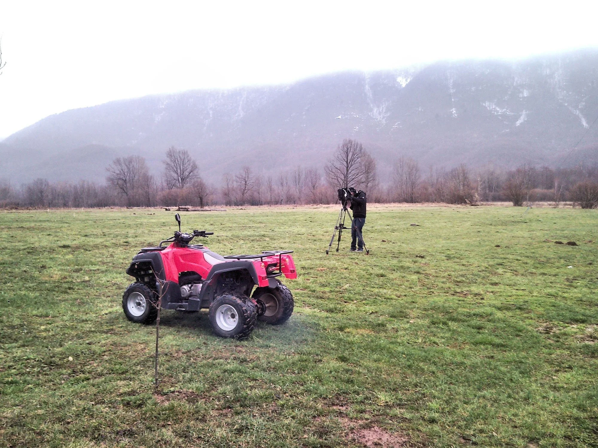 A person is filming with a camera on a tripod in an open grassy field with leafless trees, a mountain in the background, and an ATV in the foreground.