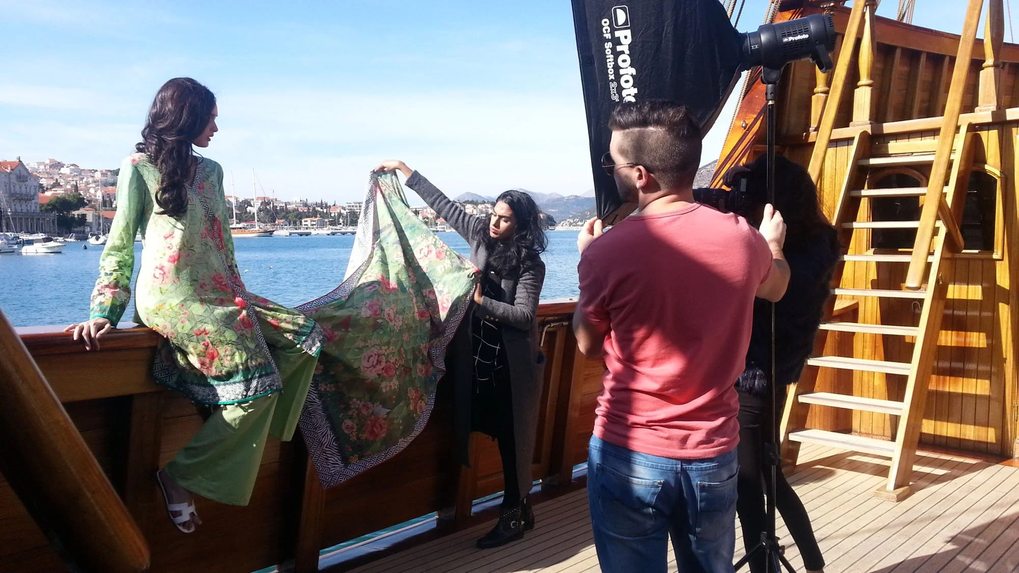 A photoshoot on a boat featuring a woman in a floral jacket and green pants, with crew members adjusting a fabric backdrop, a camera operator, and a large softbox light, overlooking a marina with boats and city buildings in the background under a cle