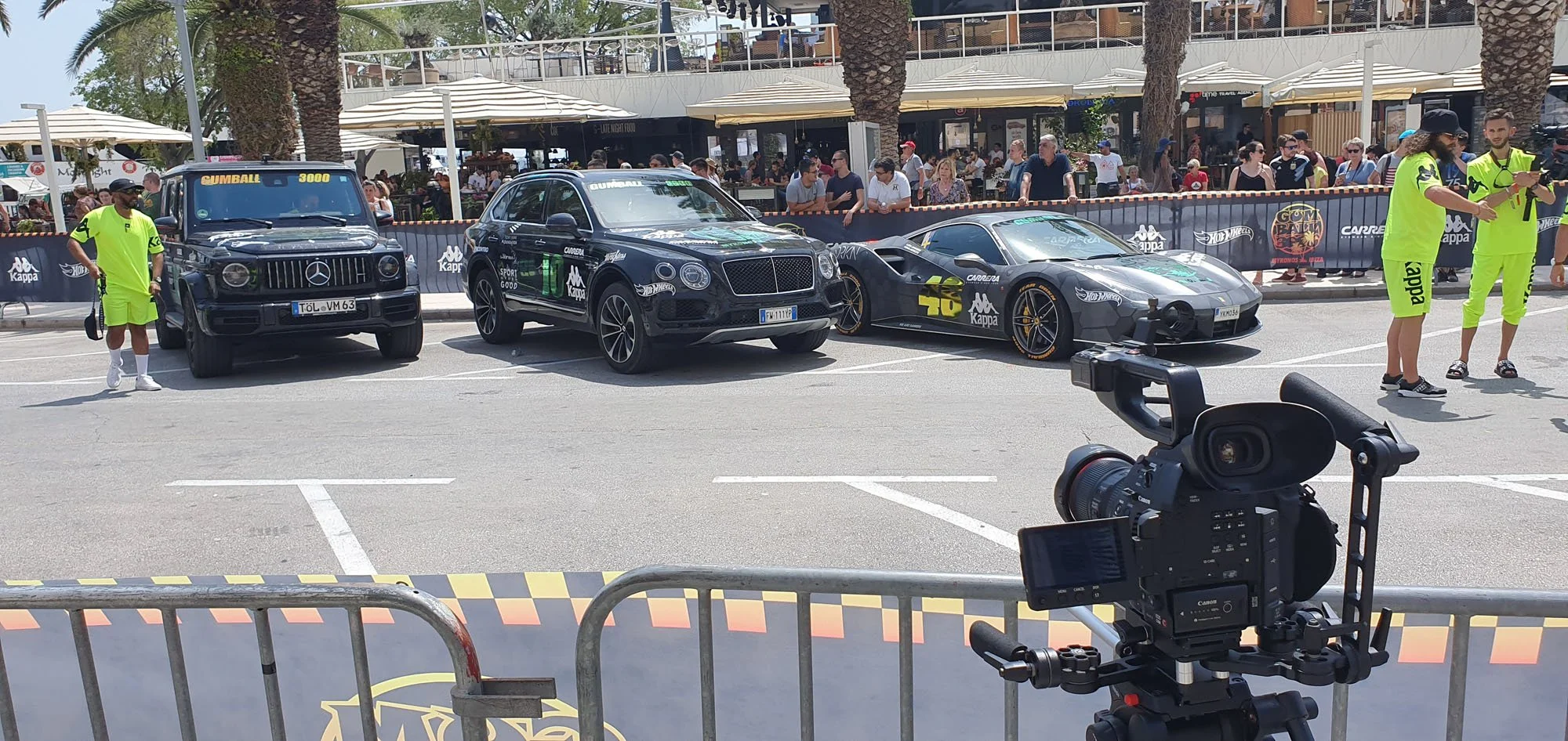 Three luxury cars, including a Mercedes-Benz G-Class, Bentley Bentayga, and Lamborghini Huracán, are parked in a row at an outdoor event with spectators behind a black barrier. Several event staff in bright yellow-green uniforms are present, with one