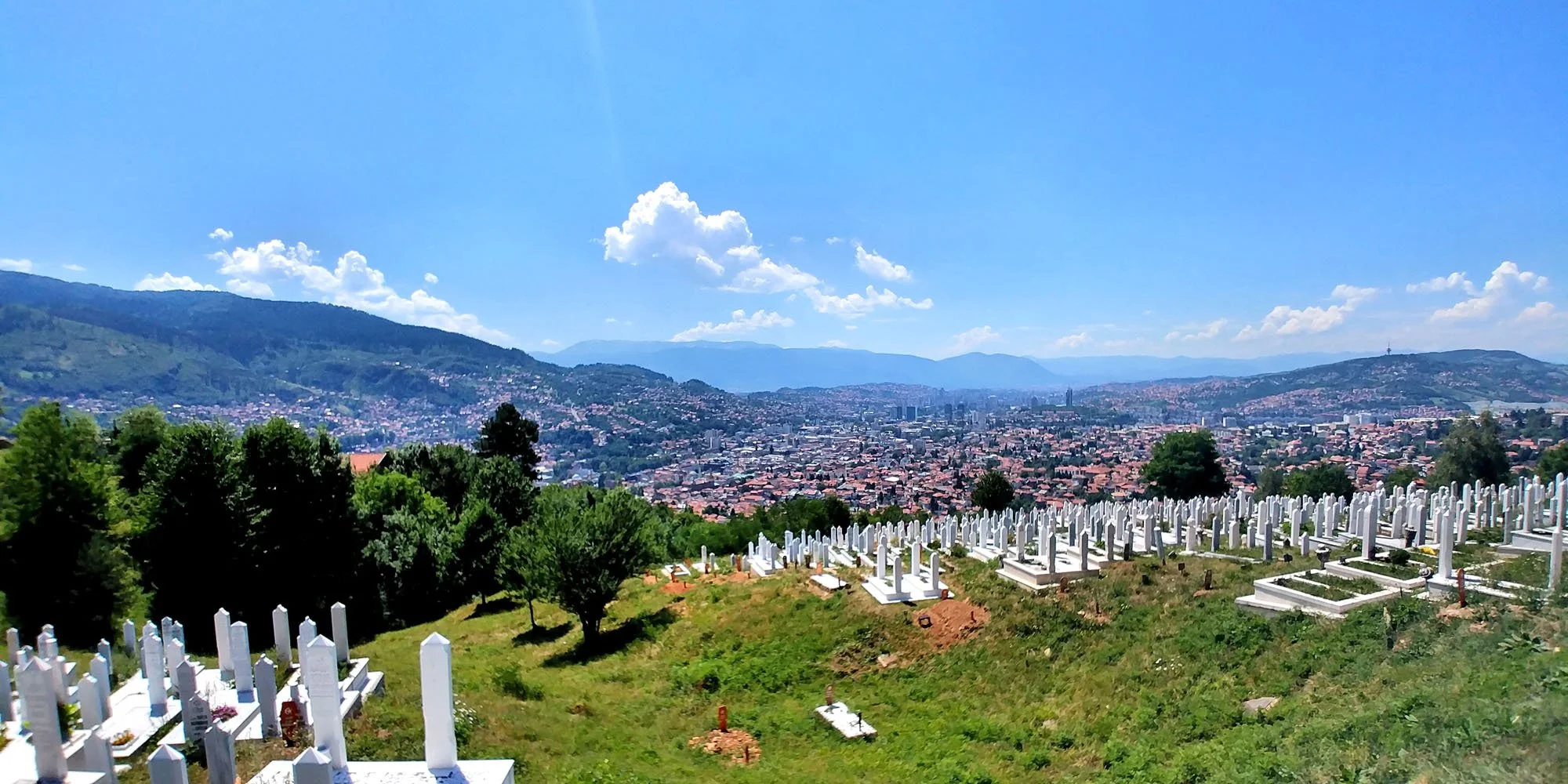 View of a cityscape with numerous buildings, surrounded by mountains and a clear blue sky with scattered clouds. In the foreground, there is a cemetery with white tombstones and green grass.