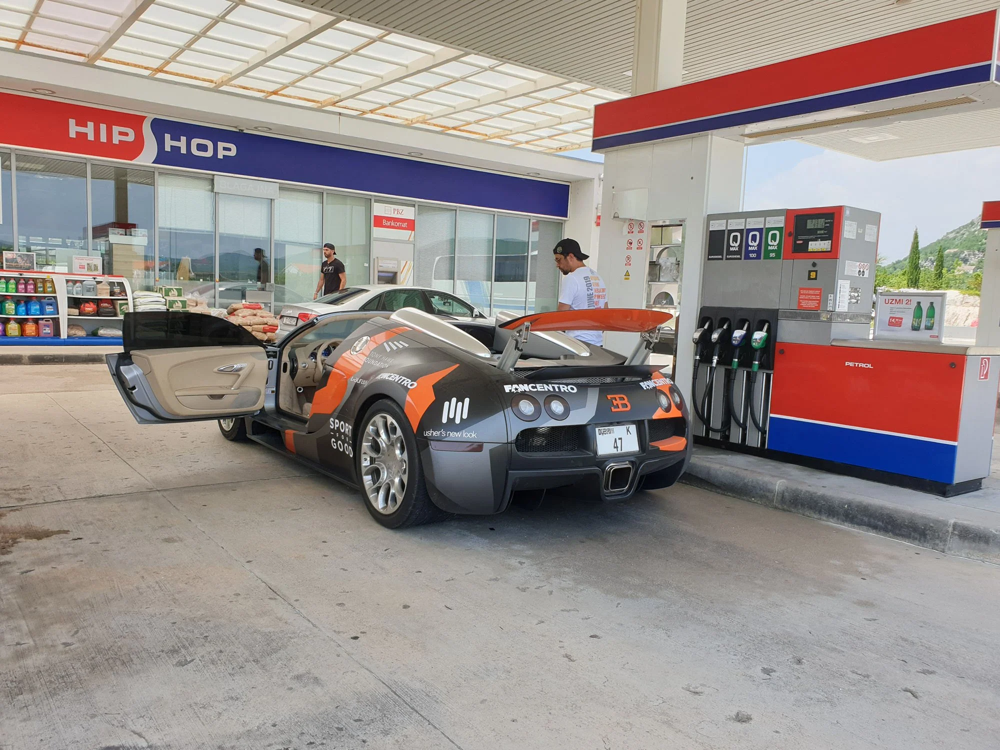A gas station with a sports car parked at the fuel pump. In the background, a convenience store labeled 'HIP HOP' is visible, with a man standing outside and another inside the store. The gas station features a covered area with a red, white, and blu
