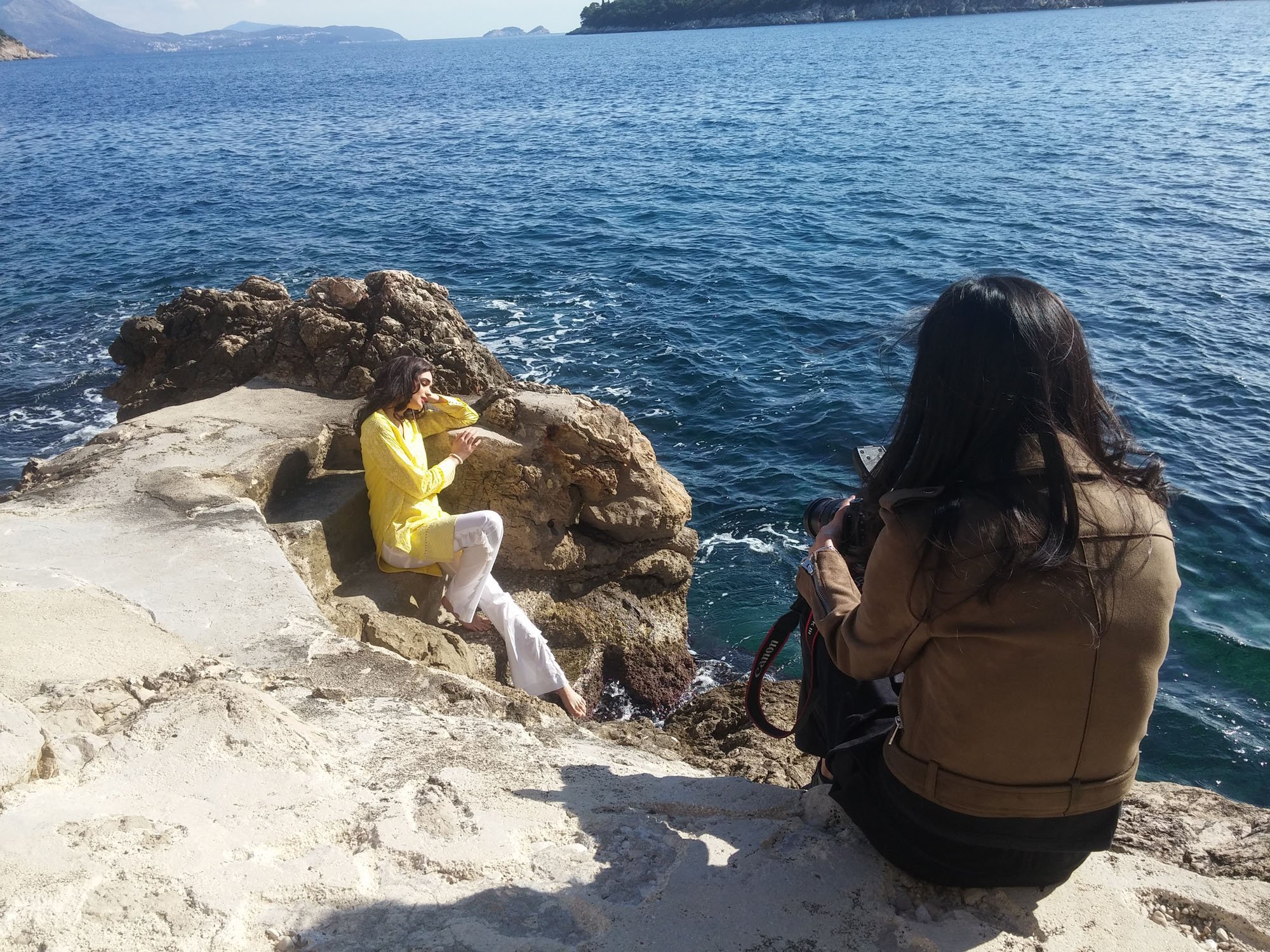 A woman wearing a yellow coat and white pants sitting on rocks near the water, while a photographer with long dark hair takes her picture. The scene is by the ocean with a boat in the distance.