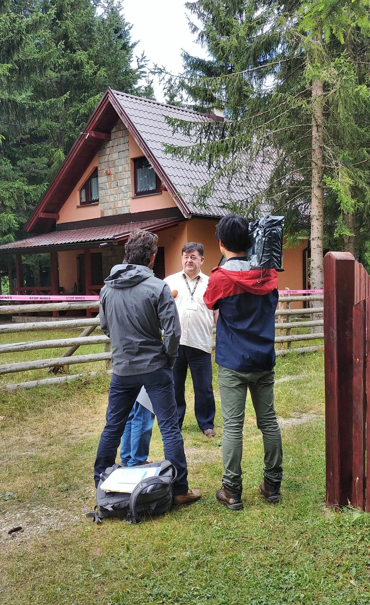 Four people are standing outdoors in front of a house surrounded by trees, engaged in conversation. One person appears to be speaking, while the others listen. There are backpacks and papers on the ground nearby.
