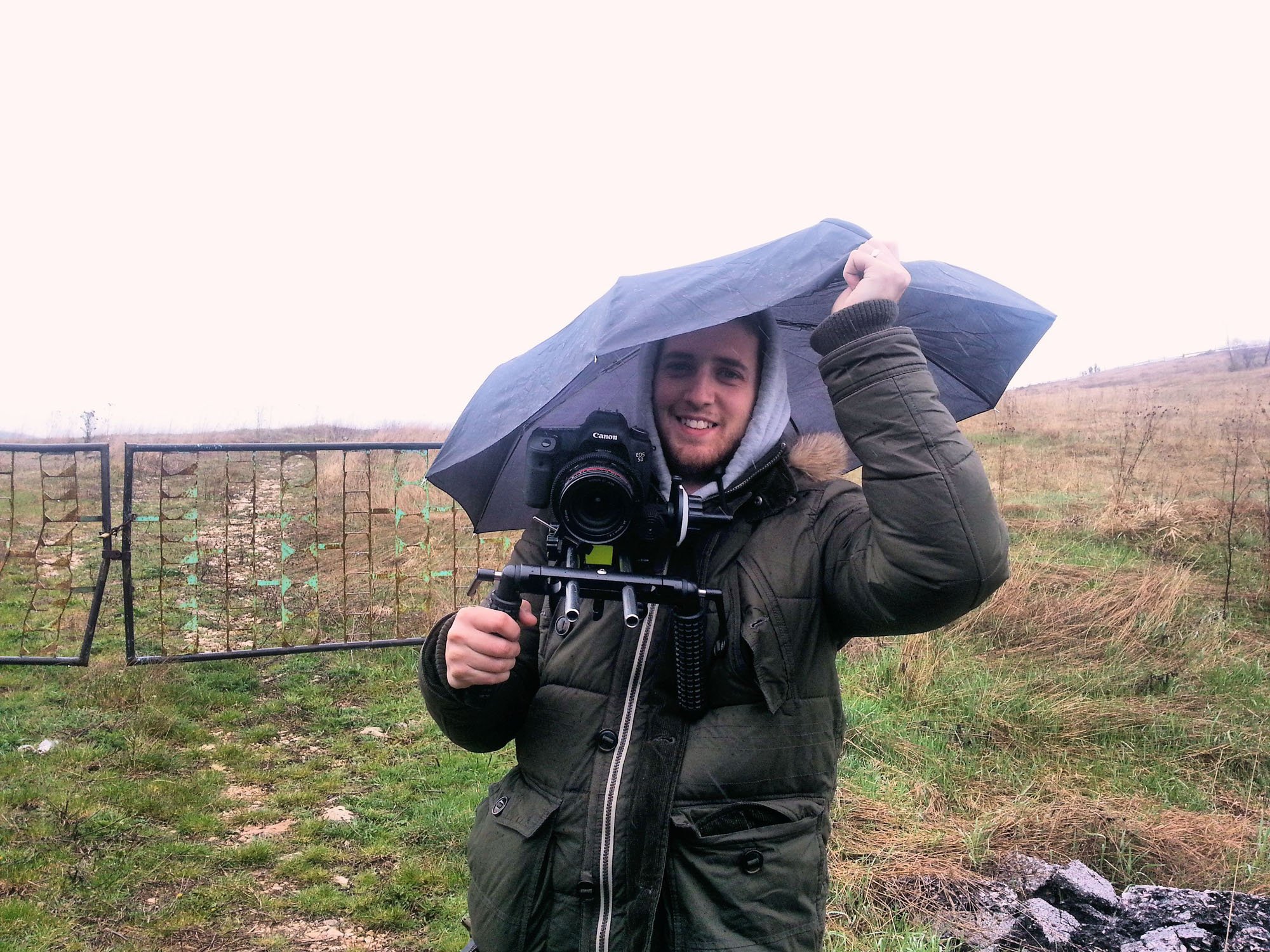 A man smiling, holding a camera with a tripod, standing outdoors in a field holding an umbrella above his head on a rainy day.