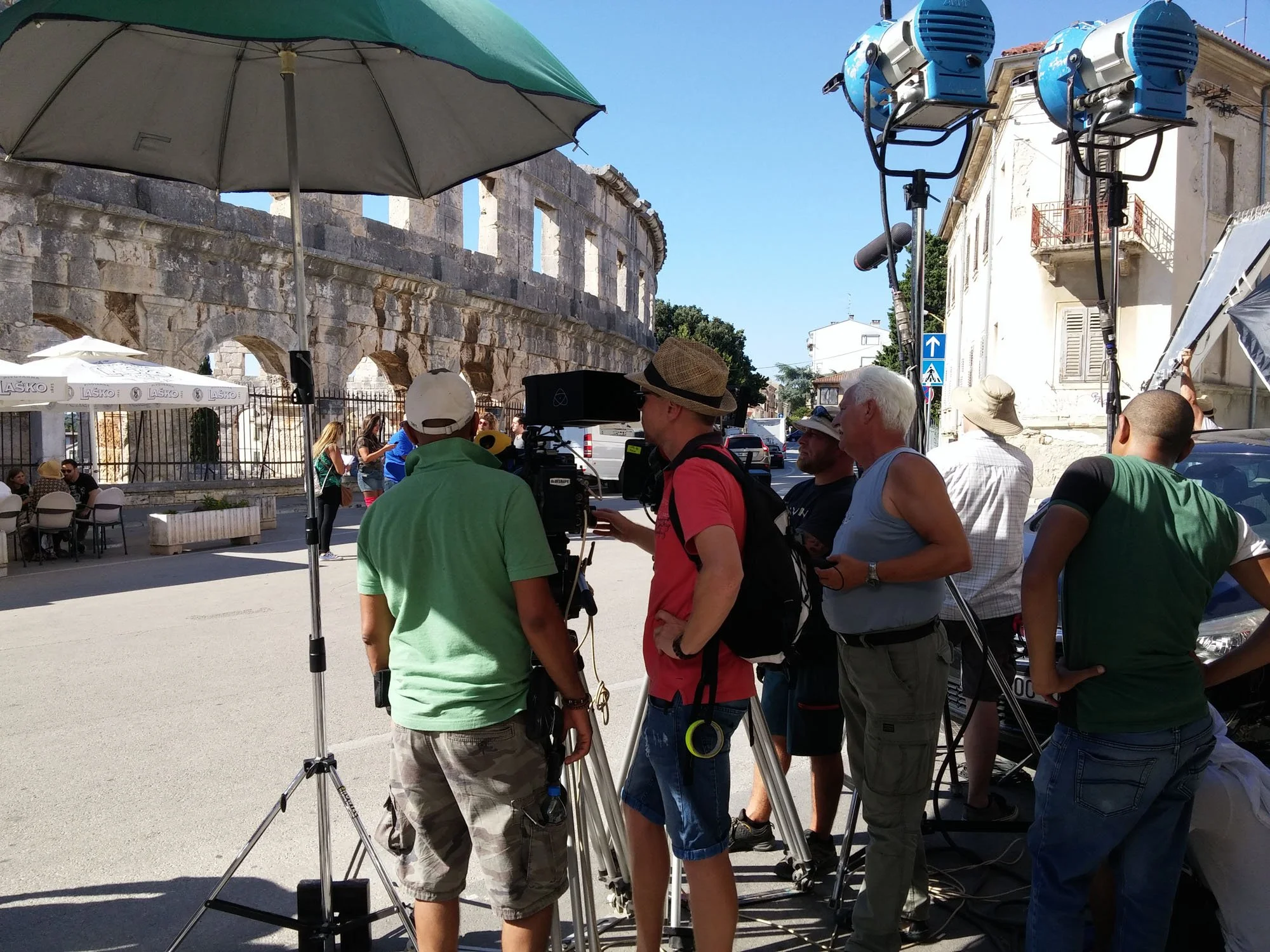 Film crew working outdoors with camera equipment in front of historic stone ruins and nearby architecture on a sunny day.