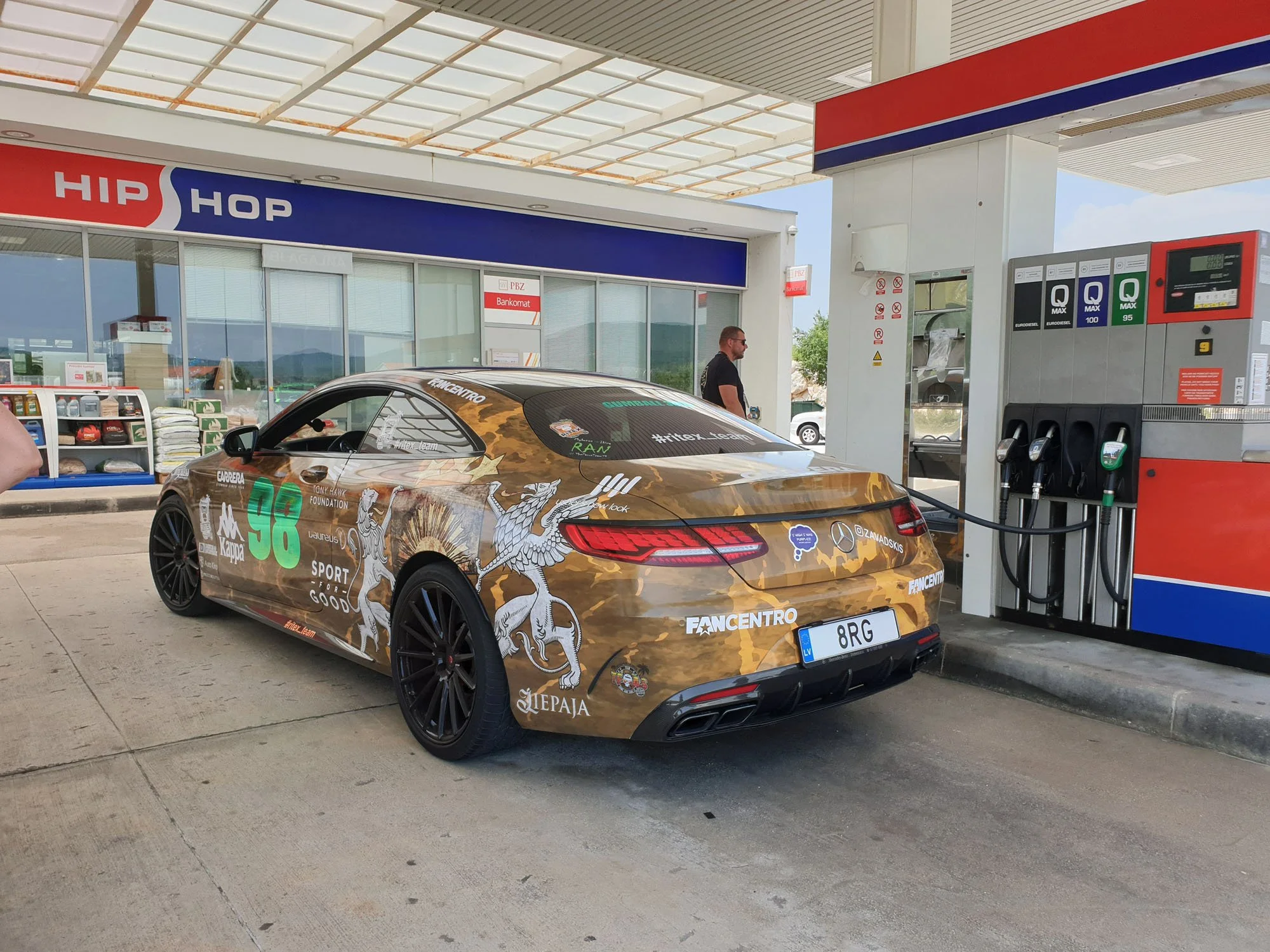 A sports car with a beige camouflage wrap featuring dragon artwork and sponsor logos parked at a gas station with a red, white, and blue sign and fuel dispensers.