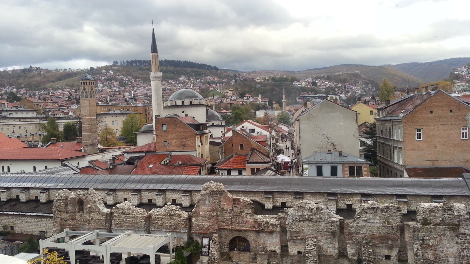 Cityscape view of a historic town with a mix of old stone ruins, church minarets, modern buildings, and distant hills under cloudy sky.