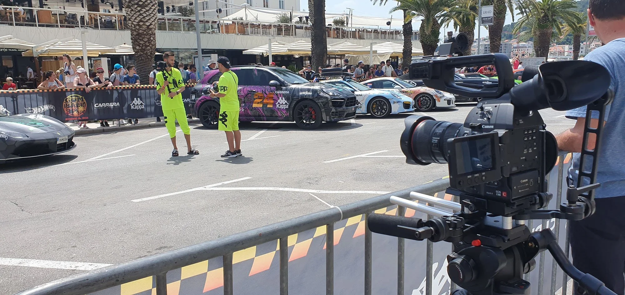 Photo of a car racing event with several high-performance cars parked in a line, spectators on the sides, and two crew members in fluorescent yellow outfits talking. There is a professional camera setup recording the scene, and a person in a gray shi