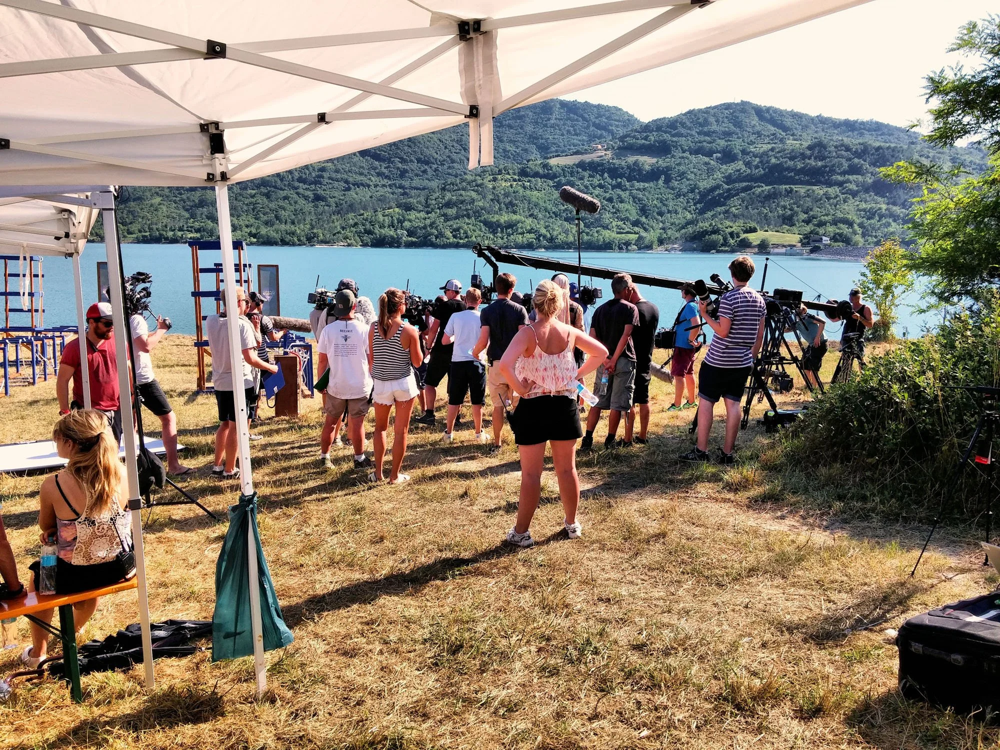 Film crew setting up equipment near a lake with mountains in the background.