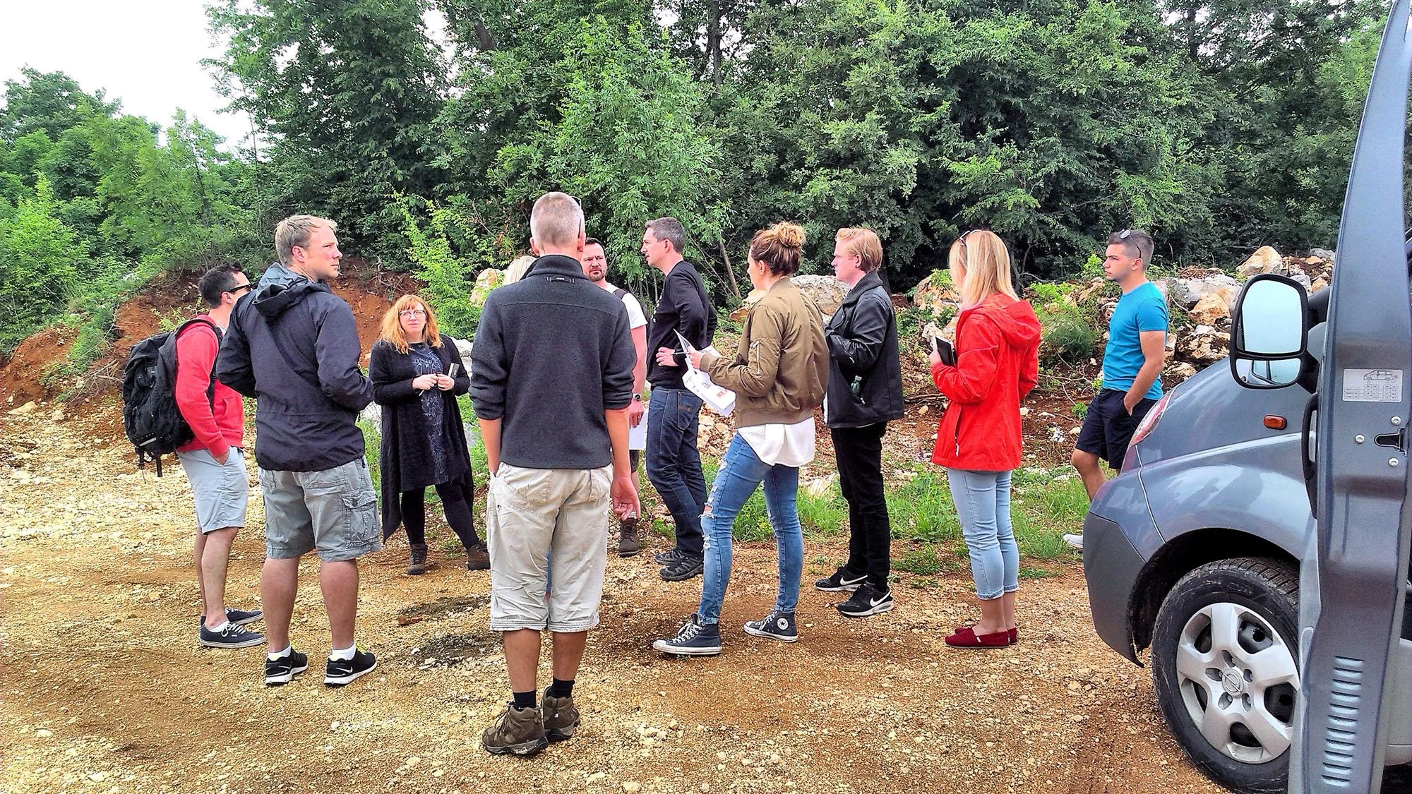 Group of people standing outdoors on a dirt ground, engaging in conversation, with trees and rocks in the background and a van parked nearby.