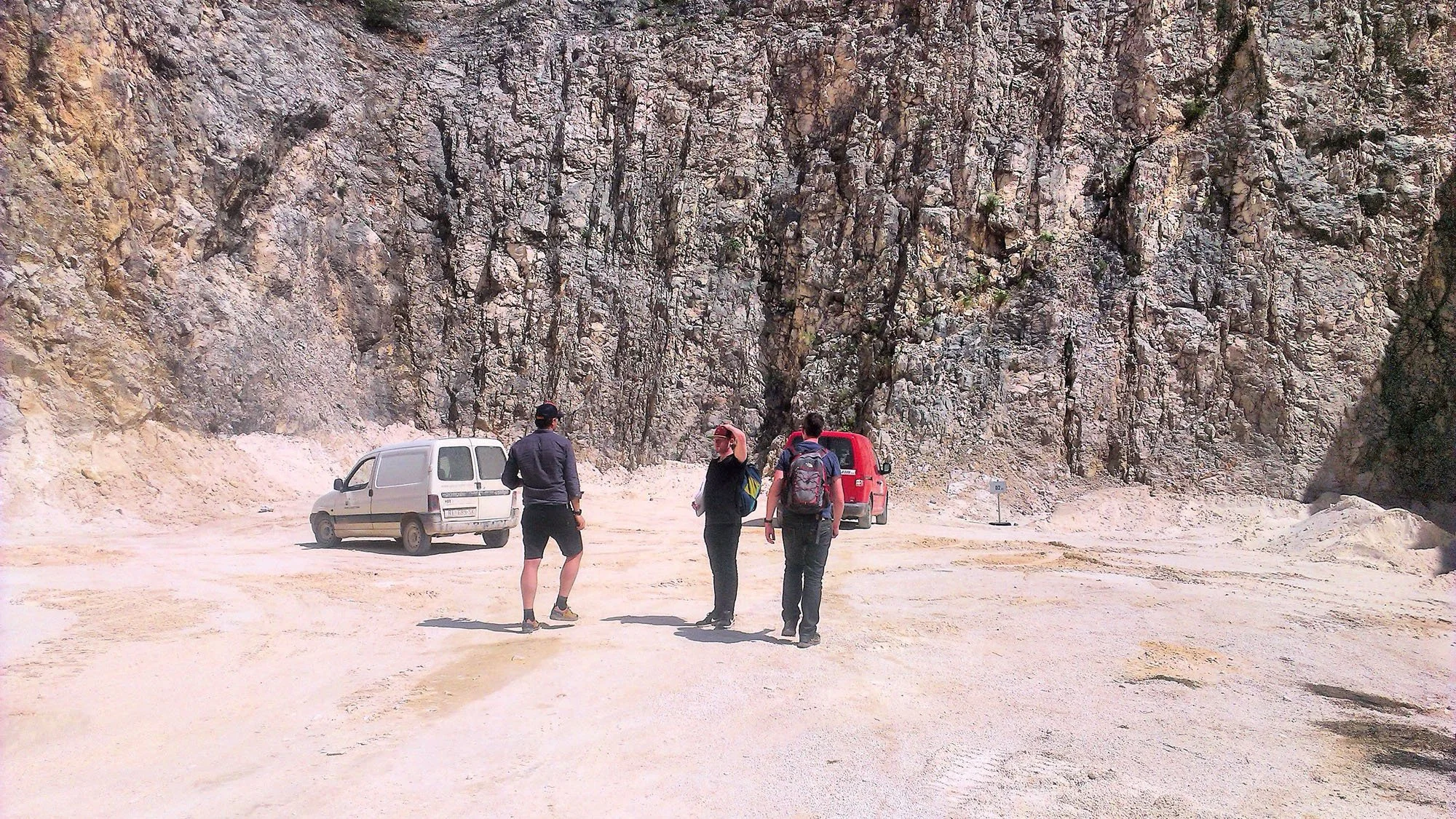 Three people standing on a dirt road in front of a rocky cliff, with two parked cars nearby.