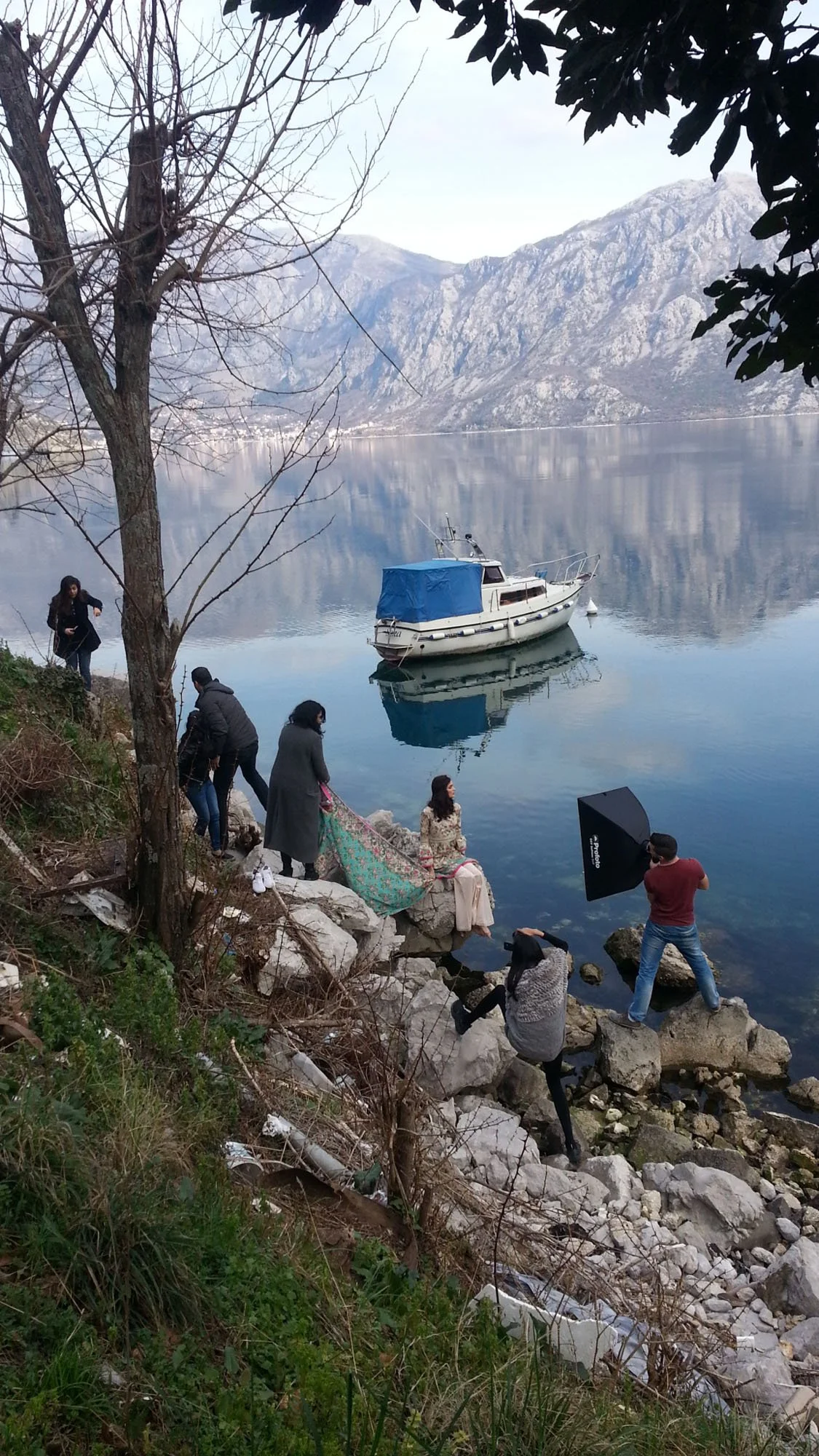 A photoshoot by a lake features a model sitting on rocks, and a photographer taking pictures with professional gear. Several people are around, some standing on the rocky shoreline, with a boat floating on the calm water and mountains in the backgrou