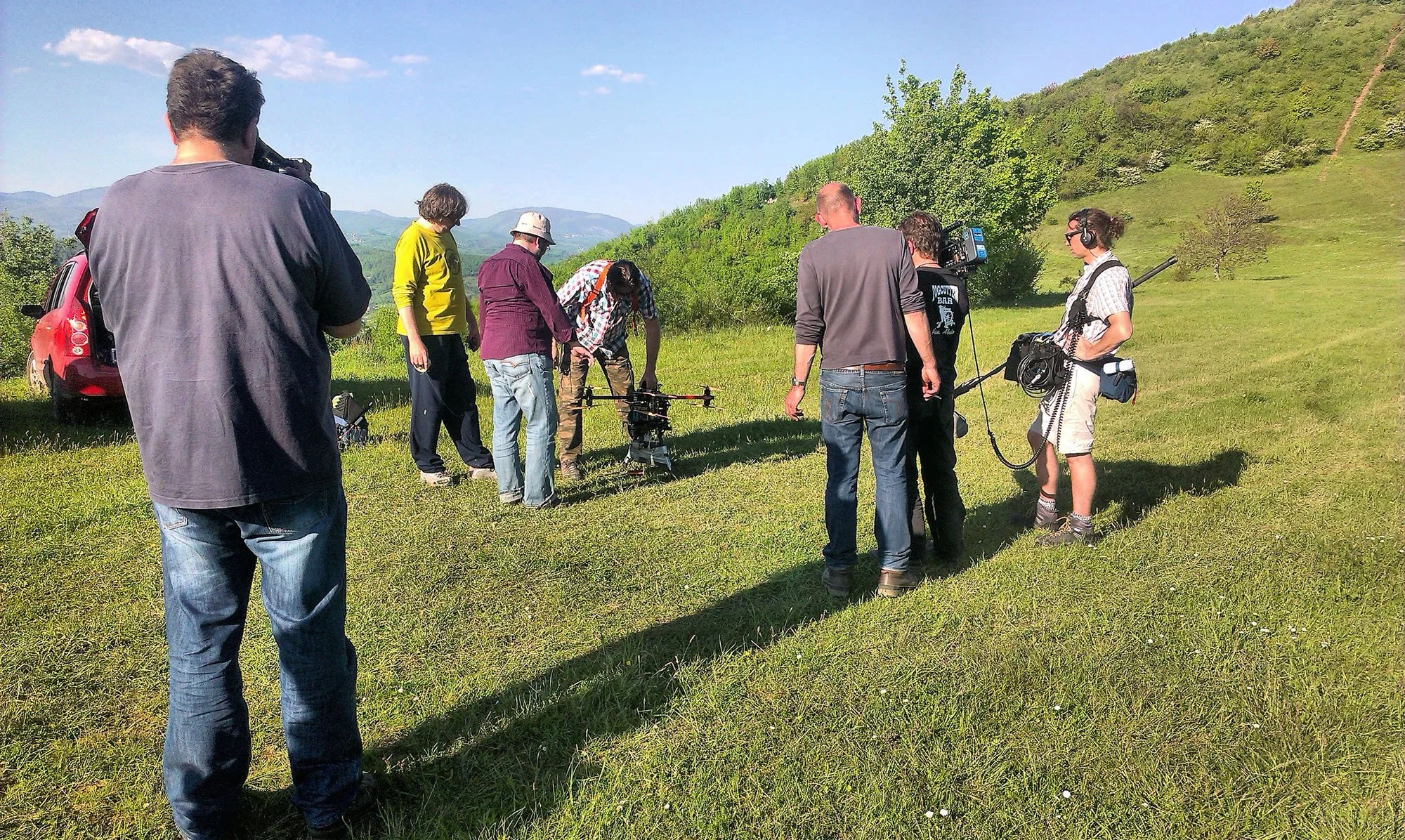 A film crew working outdoors on a grassy hillside with a scenic mountain background on a sunny day. The crew includes a man operating a drone, a woman with headphones, and several others preparing equipment.