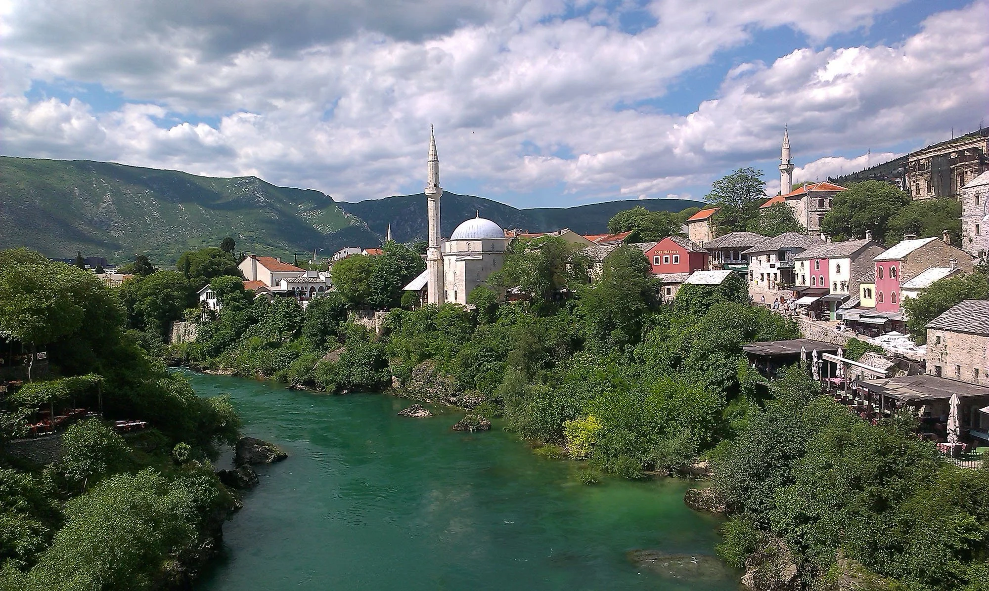 A scenic view of a river flowing through a village with colorful houses, lush green trees, and two white mosques with minarets, set against a backdrop of green mountains under a partly cloudy sky.