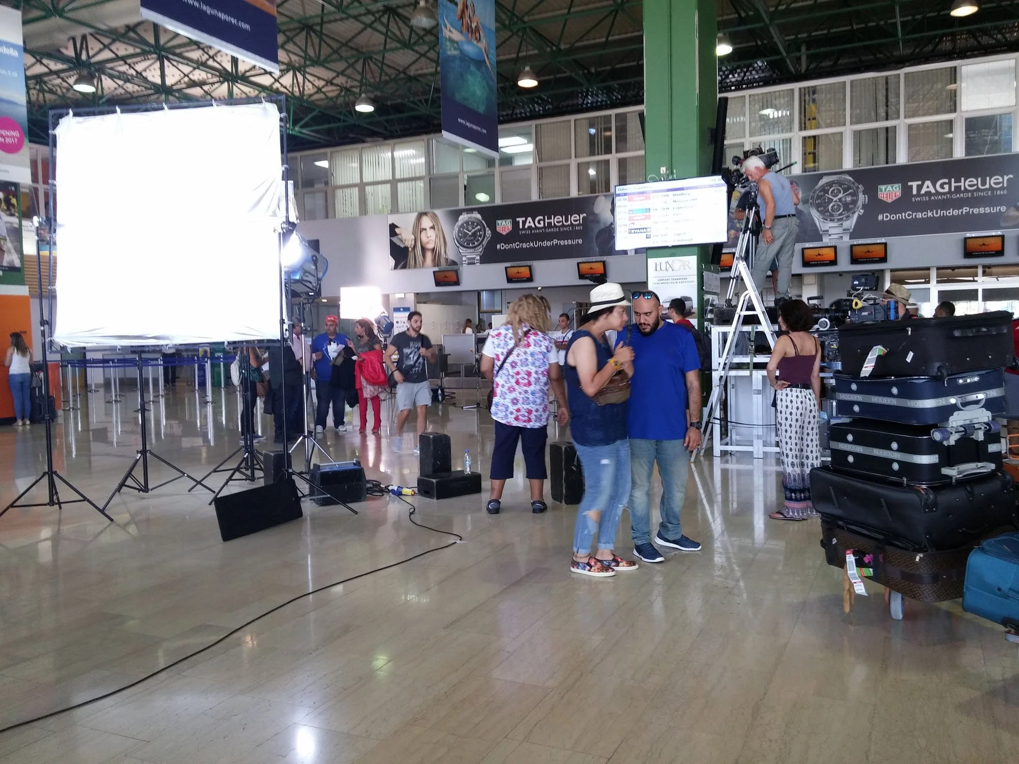People setting up filming equipment at an indoor airport or public space with advertisement banners for TAG Heuer watches.