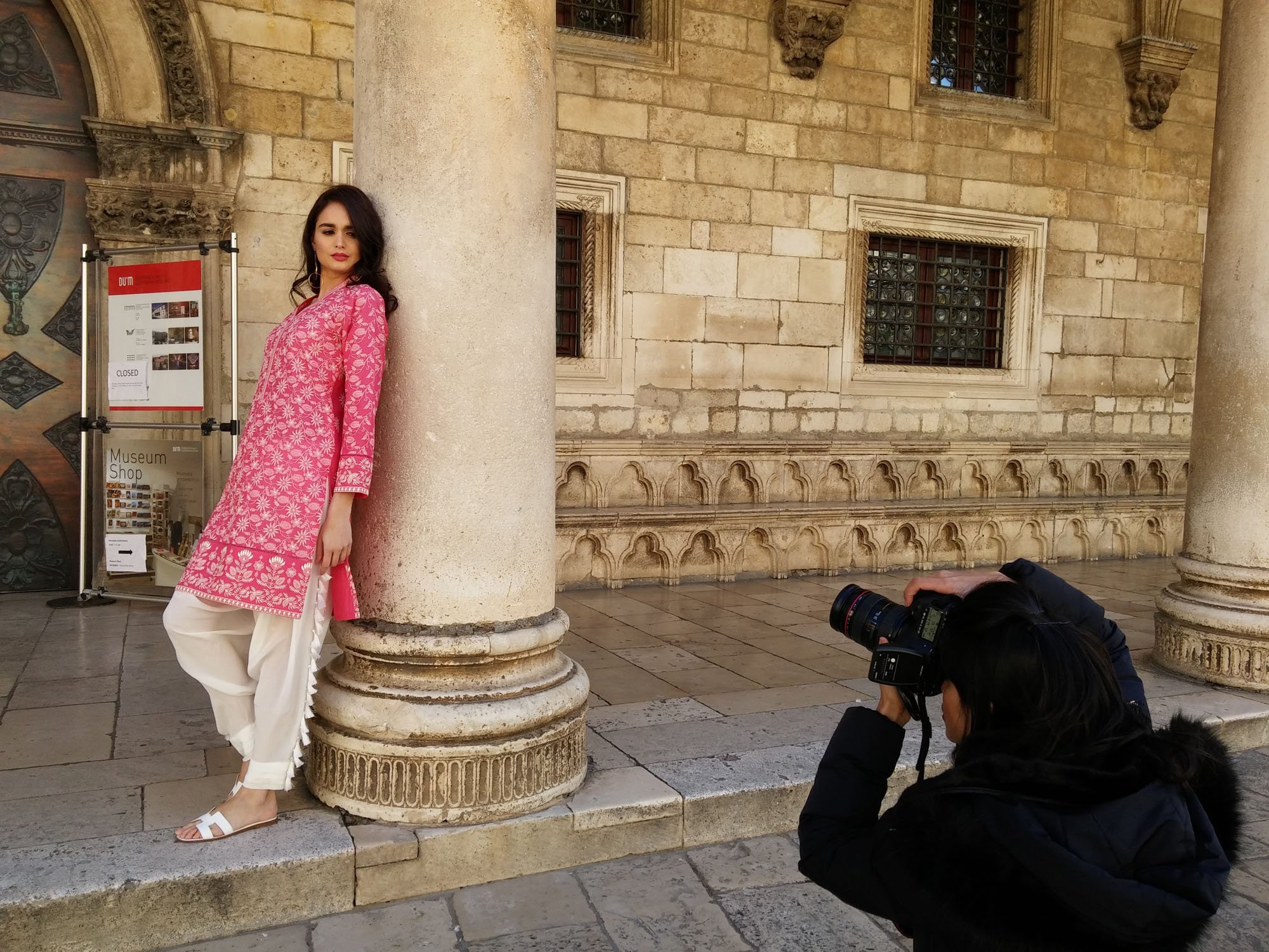 A woman in a pink traditional dress posing for a photograph at a historic building with stone columns and ornate windows. A photographer crouches with a camera to take her picture.