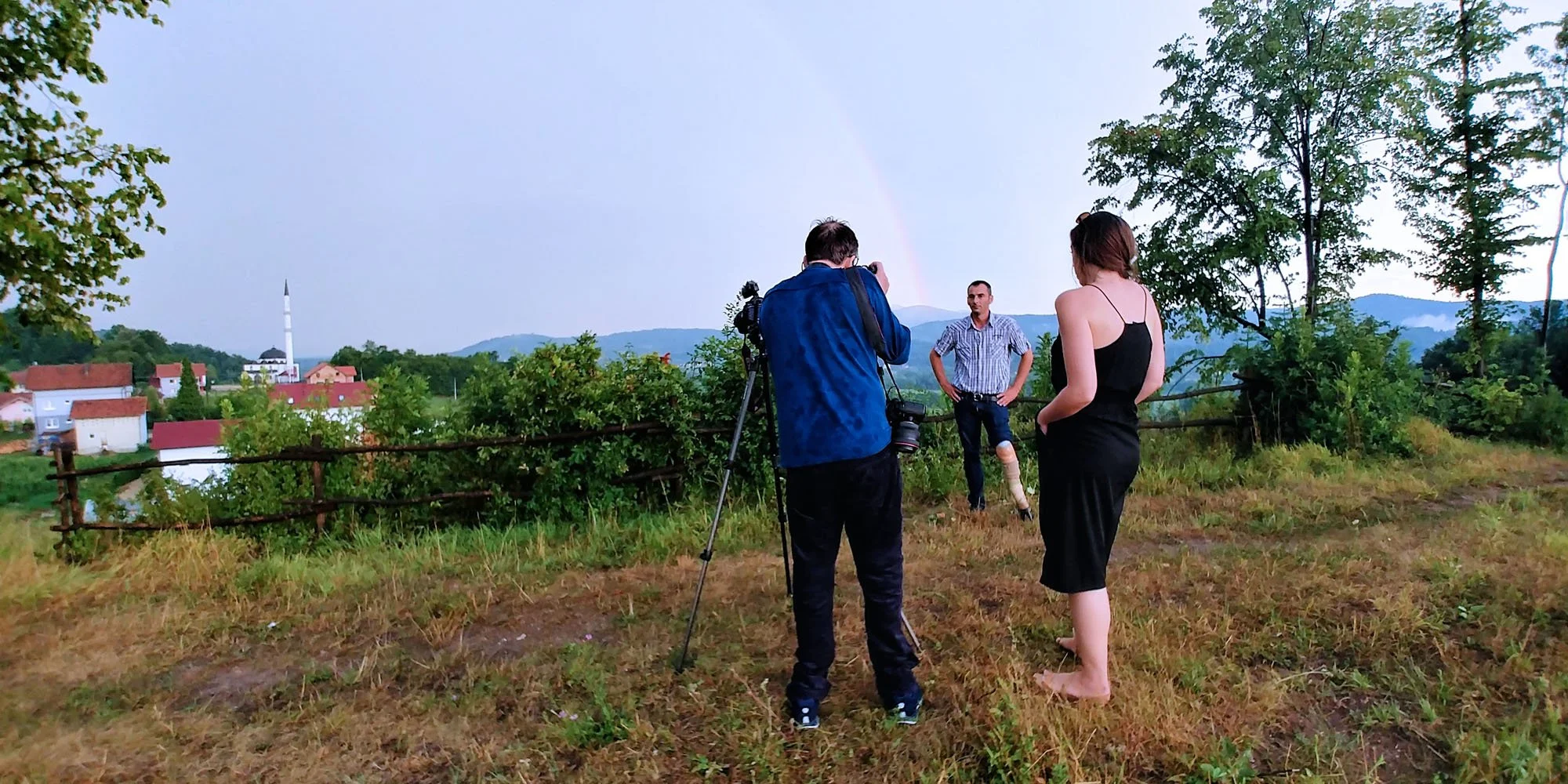 A man stands outdoors in a rural area during the daytime, with two people filming him. One person is taking a photo with a camera, and a woman with long hair in a black dress is standing nearby, facing the man. There are trees, a village with red roo