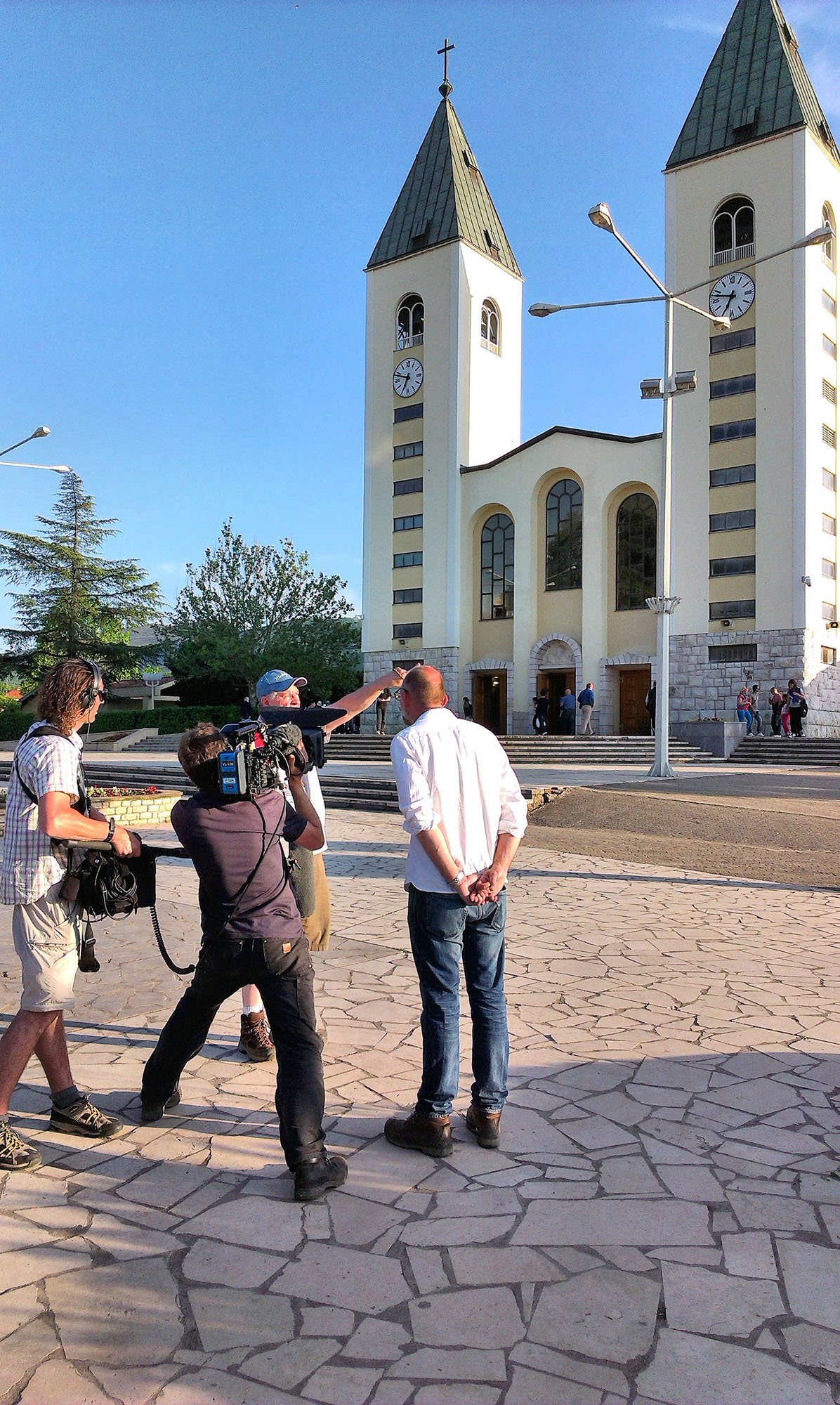 People filming a church with twin steeples and a clock on a clear day.