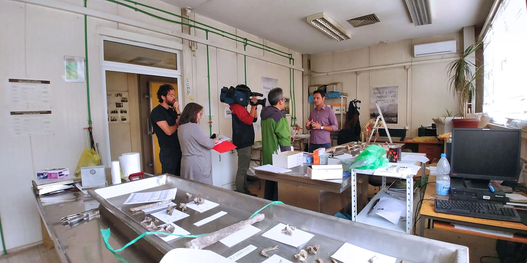 A group of people, including a cameraman, are standing in a cluttered office or research room, engaged in a discussion. The room contains desks with scattered papers, boxes, a computer monitor, a water bottle, and plants. The walls have posters, and 